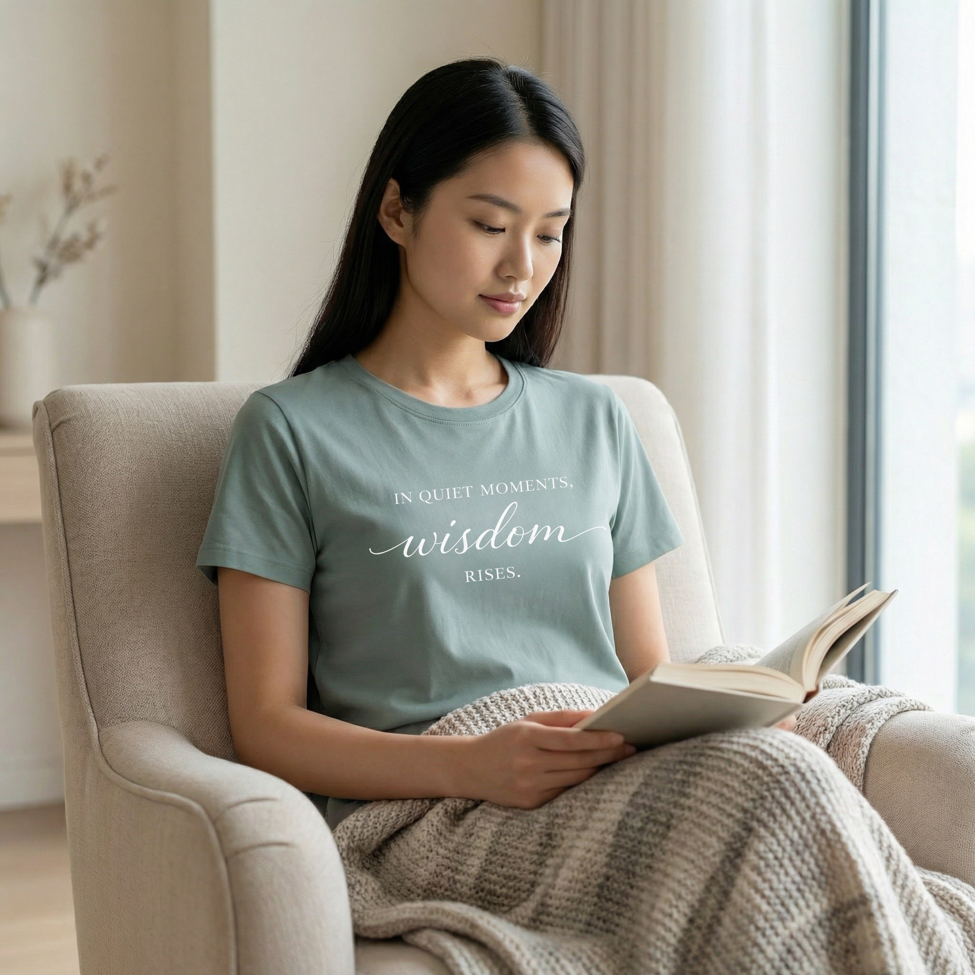Woman wearing a sage green inspirational graphic tee with the quote “In quiet moments, wisdom rises.” seated indoors reading a book in soft natural light with a calm neutral interior and elevated feminine wellness styling.