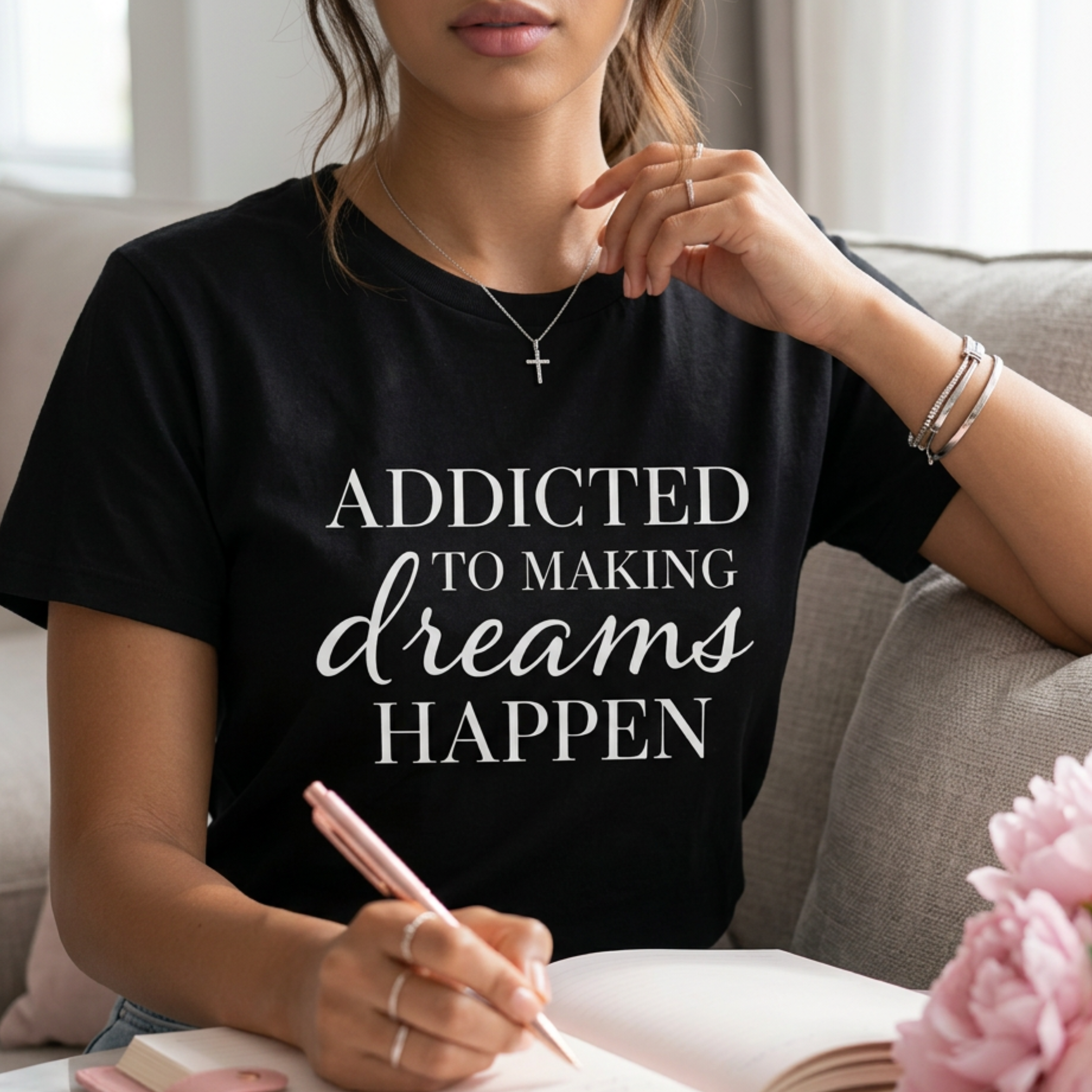 Black women’s inspirational graphic tee with “Addicted to Making Dreams Happen” slogan, styled in a bright modern interior with city views, minimal luxury aesthetic, and soft natural light. Entrepreneur woman is journaling.
