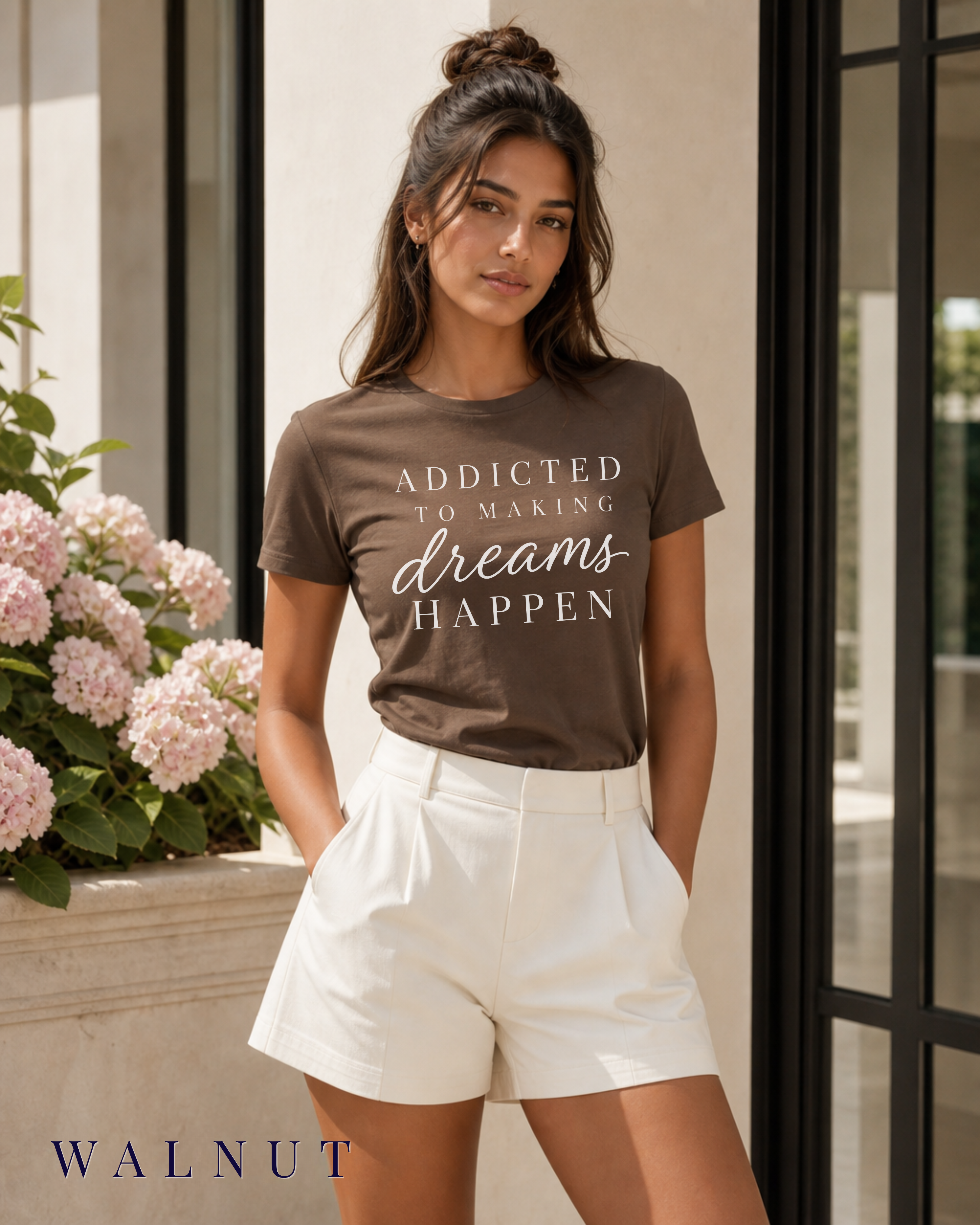 A woman wearing a brown graphic t-shirt with the text 'ADDICTED TO MAKING dreams HAPPEN' in white, paired with white shorts, standing in front of a modern building with pink hydrangeas.