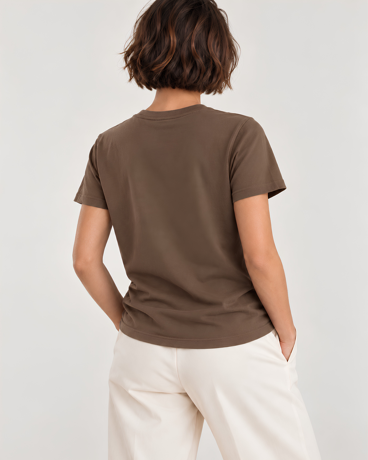 A woman shown from the back wearing a short-sleeve crew-neck t-shirt in a solid medium brown color, paired with off-white trousers, against a plain light gray studio background.