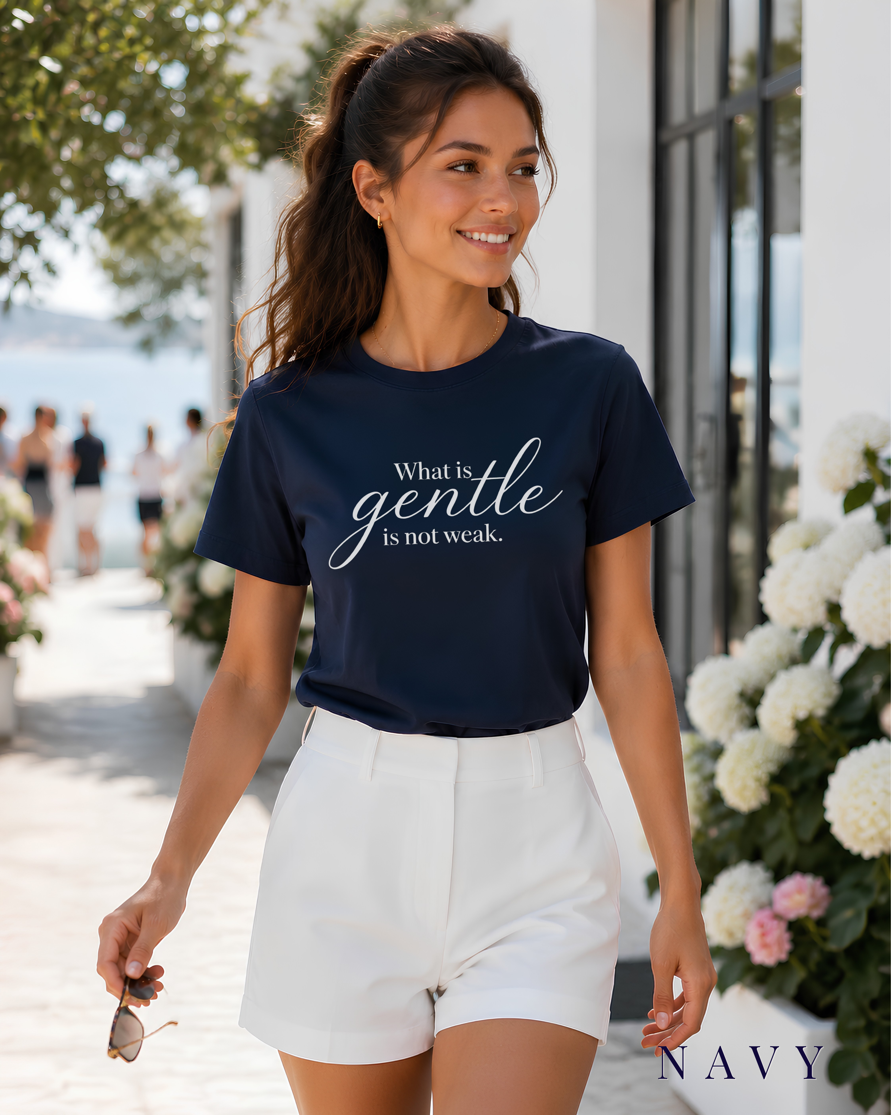 A woman walking outdoors wearing a navy Christian graphic T-shirt with the text 'What is gentle is not weak.' in white script, paired with white shorts.