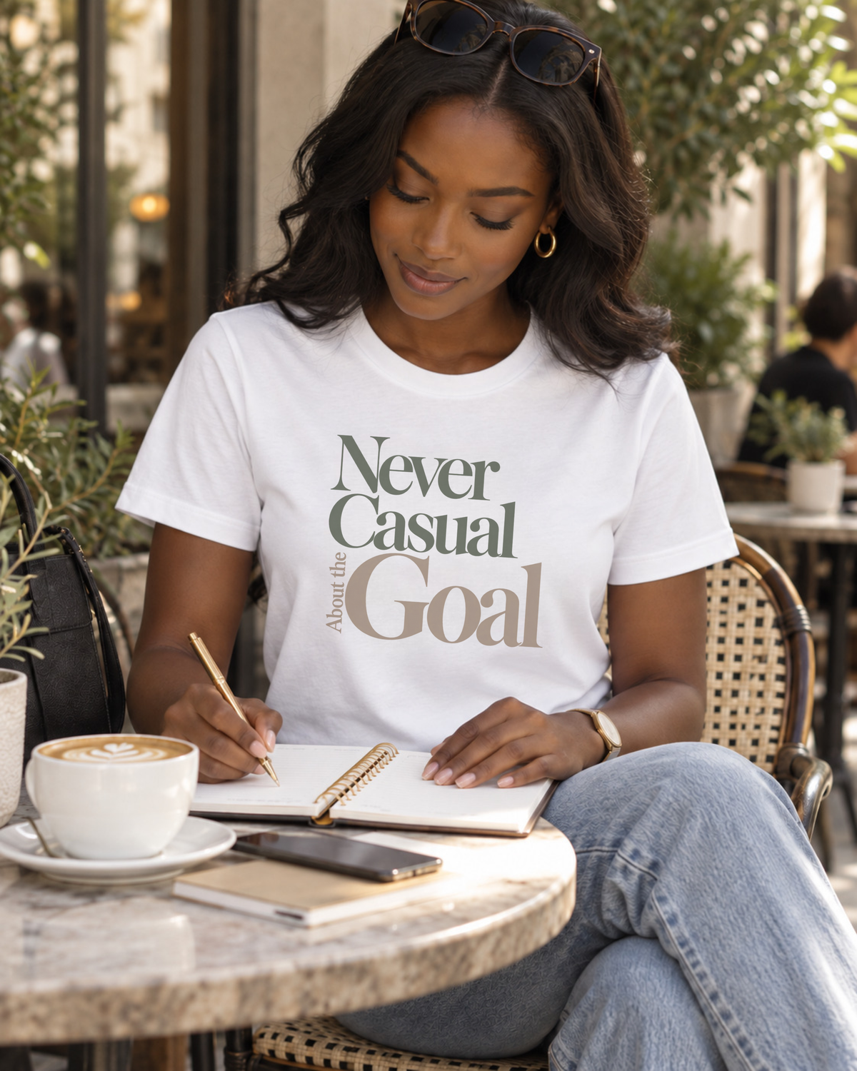 A Black woman sitting at an outdoor cafe table wearing a white motivational graphic t-shirt with the text 'Never Casual About the Goal' in green and brown lettering. She is writing in a notebook with a gold pen, with a cup of coffee, a phone, and a notebook on the table in front of her.