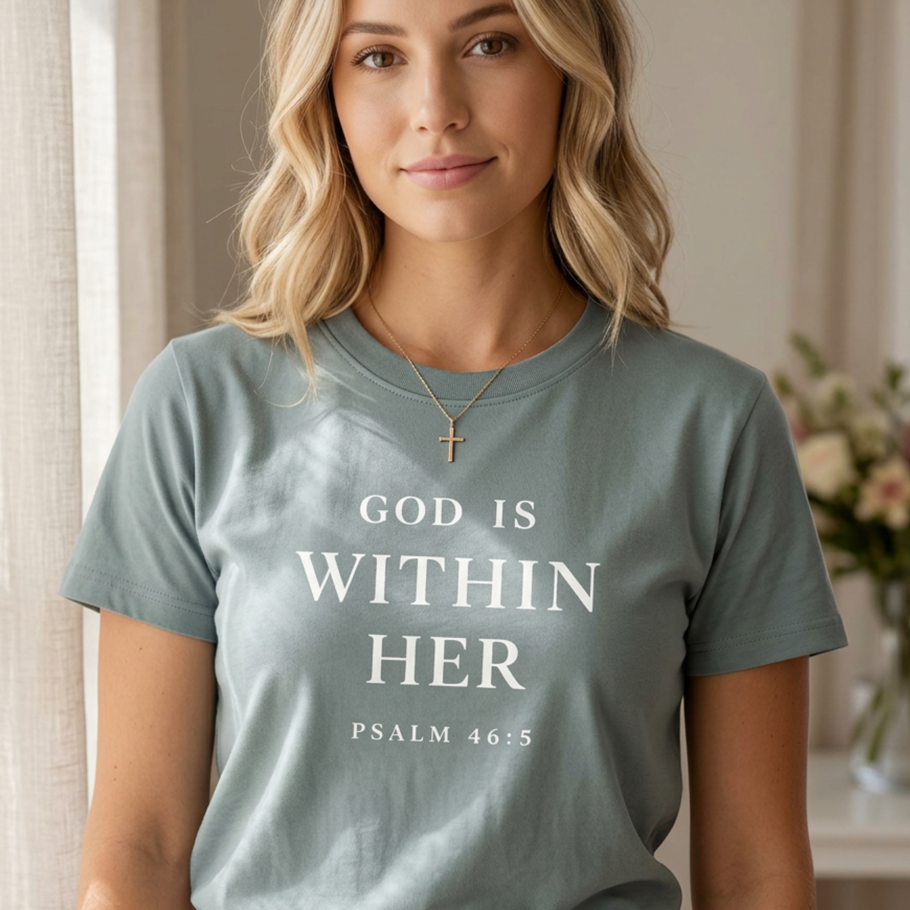 God is within her shirt in mineral color. Christian woman is standing in sunlight next to a window with flowers behind her. Bible quote verse on her tee.