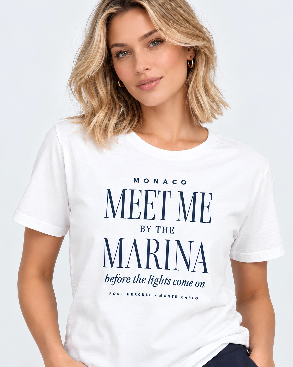 A woman wearing a white short-sleeve graphic T-shirt with navy blue text that reads 'MONACO MEET ME BY THE MARINA before the lights come on' on a light gray studio background.