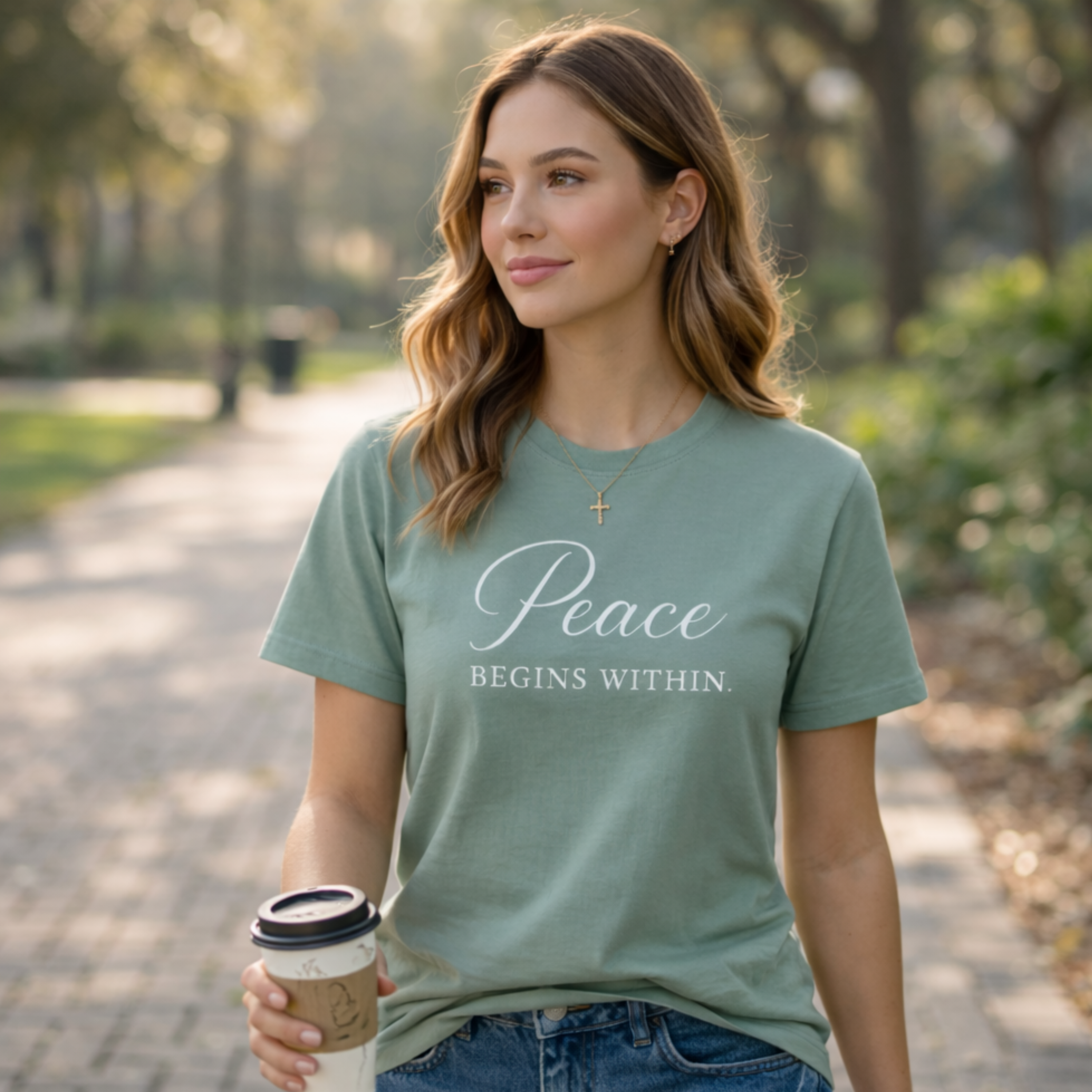 Woman wearing a sage green inspirational graphic tee with the quote “Peace Begins Within,” walking outdoors with a coffee in soft natural light, styled with jeans and a feminine self-care lifestyle aesthetic.