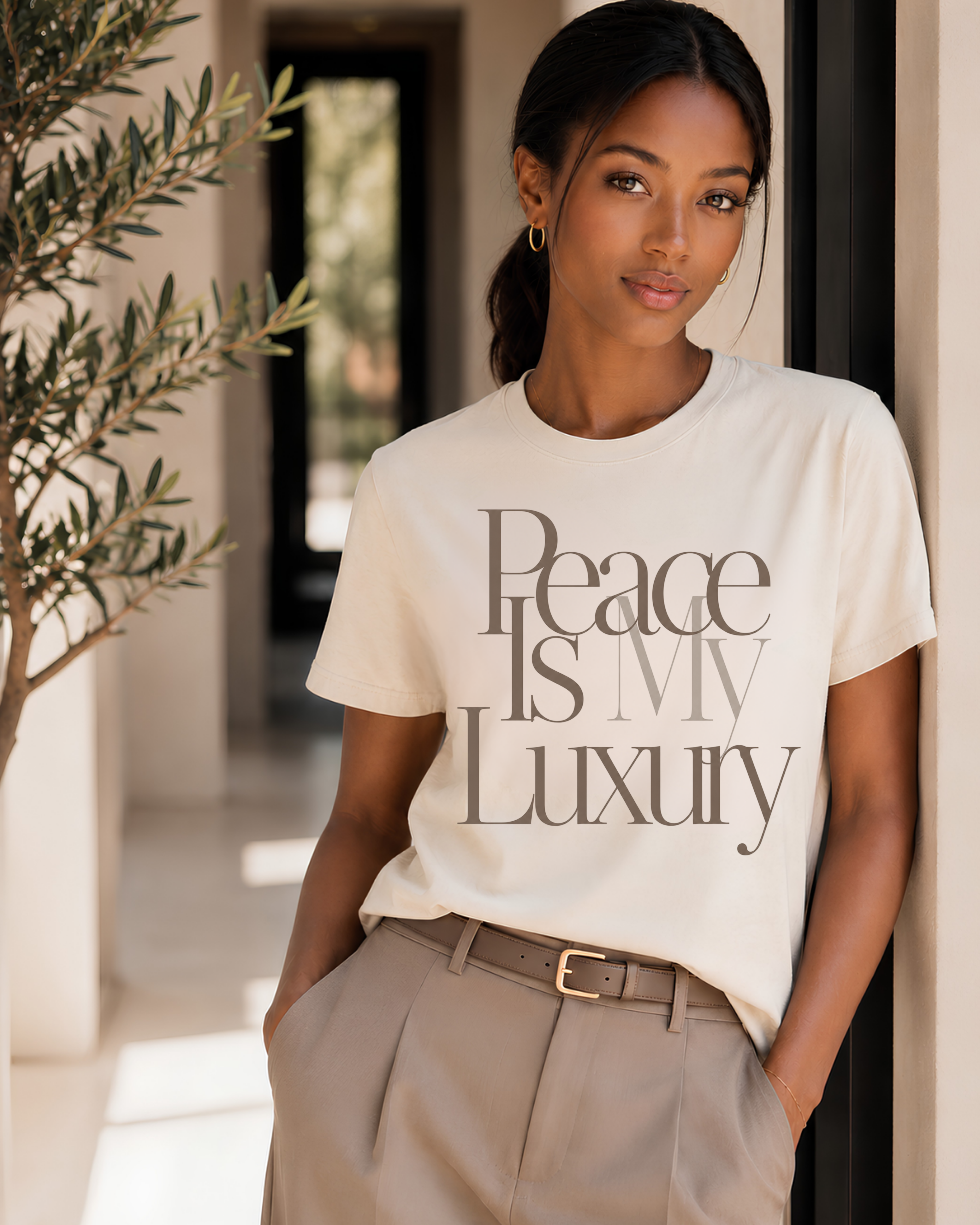Black woman wearing a bone Peace Is My Luxury T-shirt with taupe tailored trousers in a bright luxury interior, styled as a refined women’s graphic tee.
