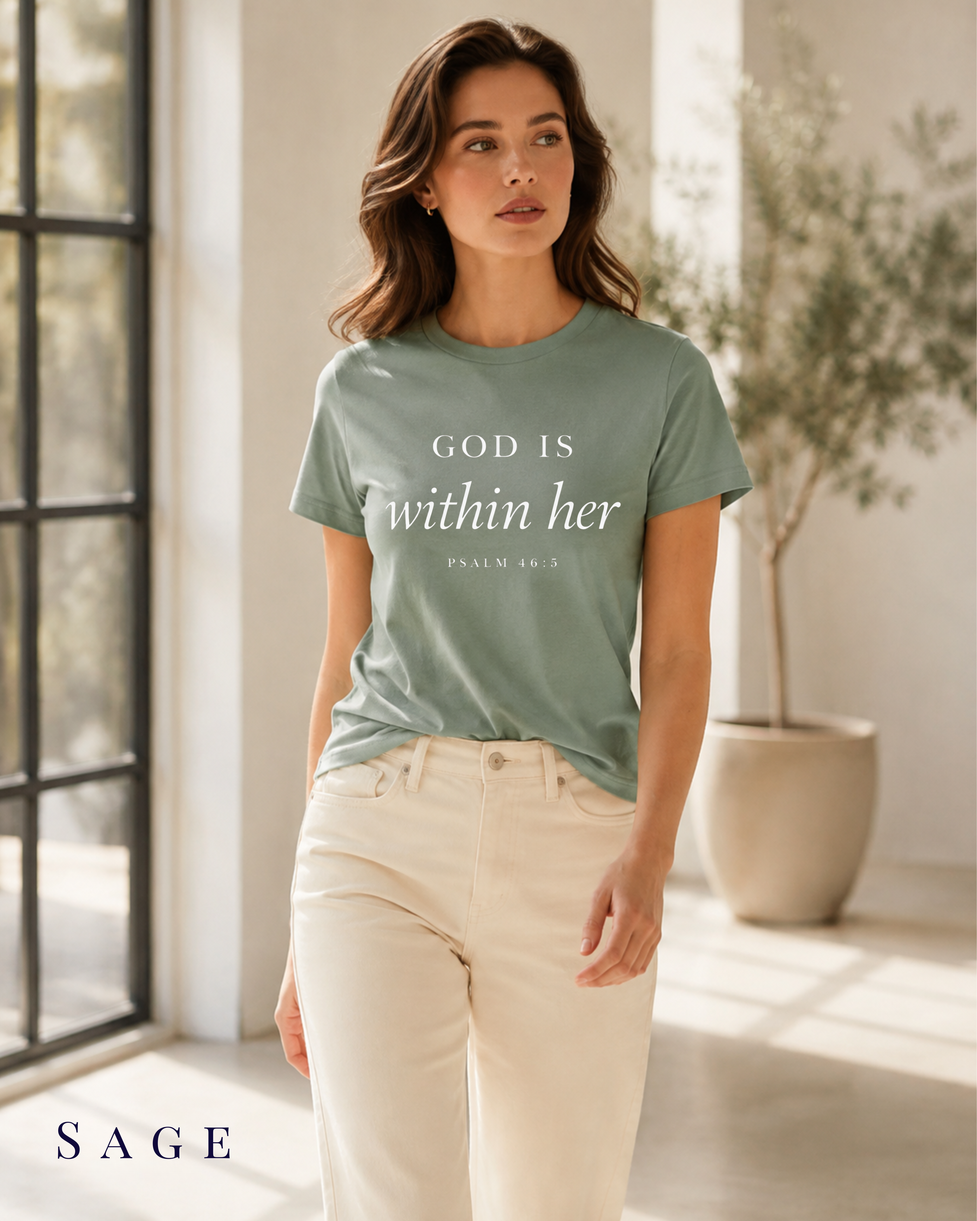 A woman wearing a sage green graphic t-shirt with the text 'GOD IS within her' and 'PSALM 81:3' printed in white, paired with cream-colored pants, standing in a bright, modern room with a window and potted plant. The color label 'SAGE' is visible in the bottom left corner.