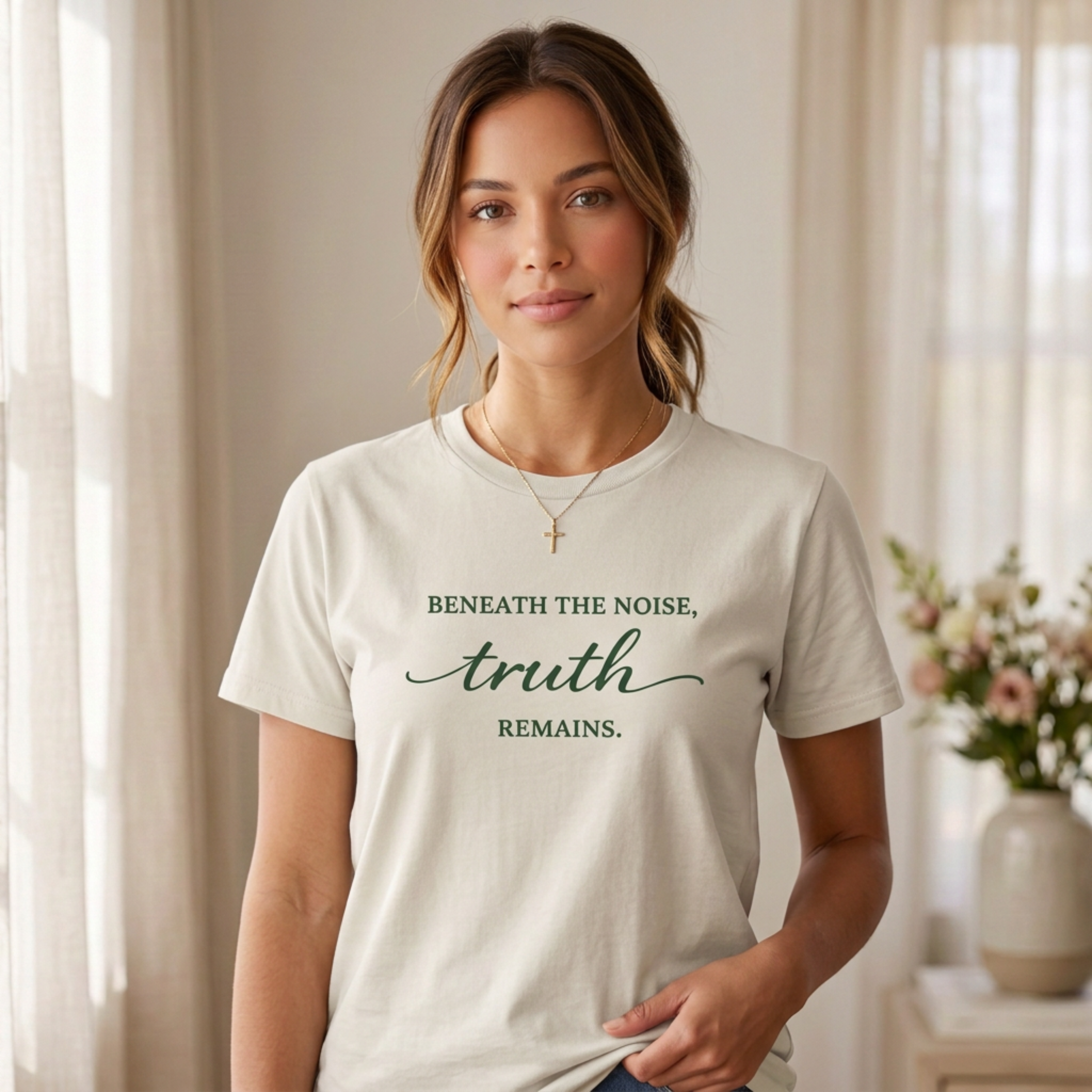 Woman wearing a bone inspirational graphic tee with the quote “Beneath the noise, truth remains.” photographed in a soft neutral interior with natural light, delicate floral accents, and an elevated feminine wellness lifestyle aesthetic.
