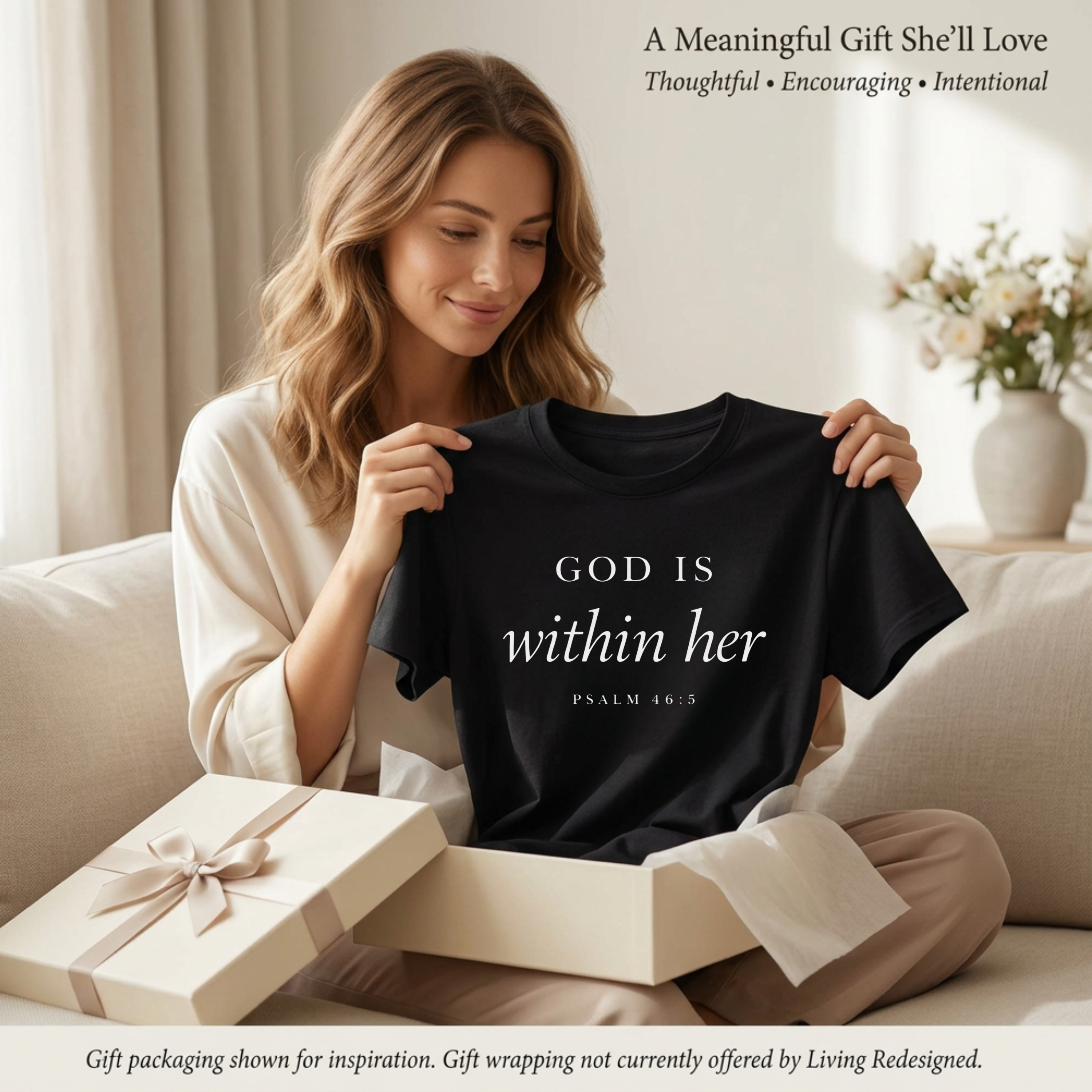 Gift for her of a scripture tee. A woman is holding up a God is within her Psalm 46:5 bible verse on a black T-shirt that she received as a gift. She is pulling it out of the gift box. There are flowers behind her.