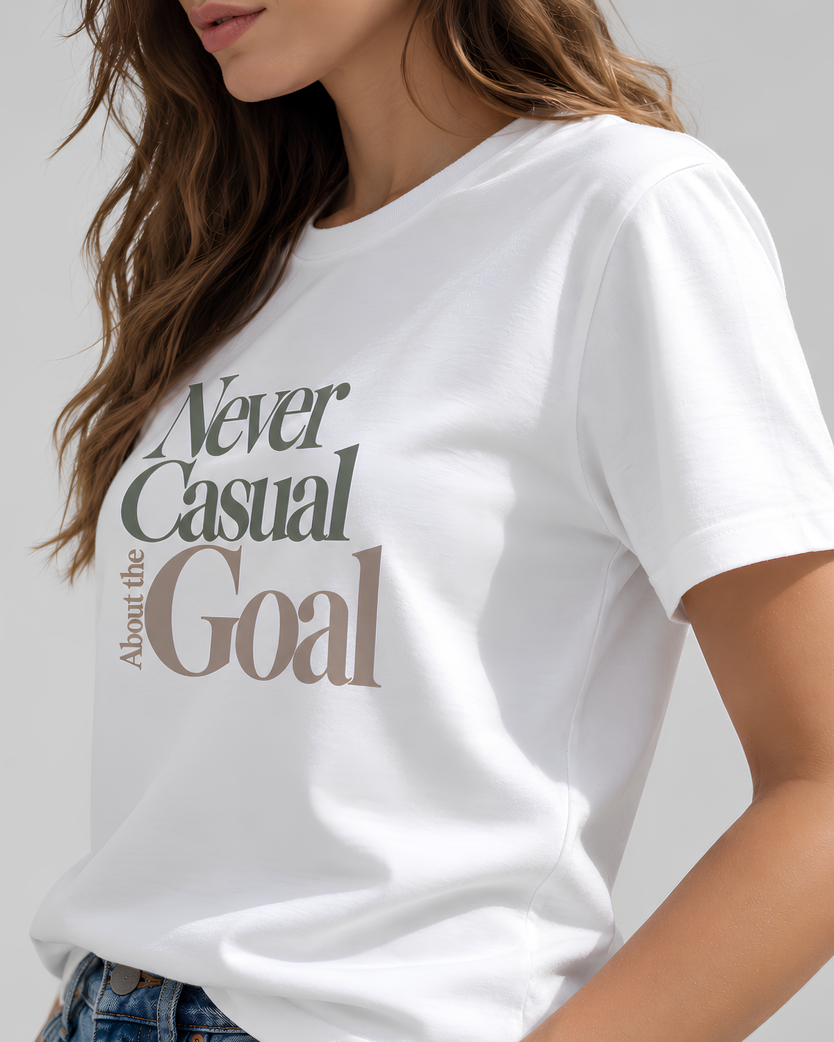 Close-up of Never Casual About the Goal graphic T-shirt showing olive-gray and taupe lettering printed on white cotton.