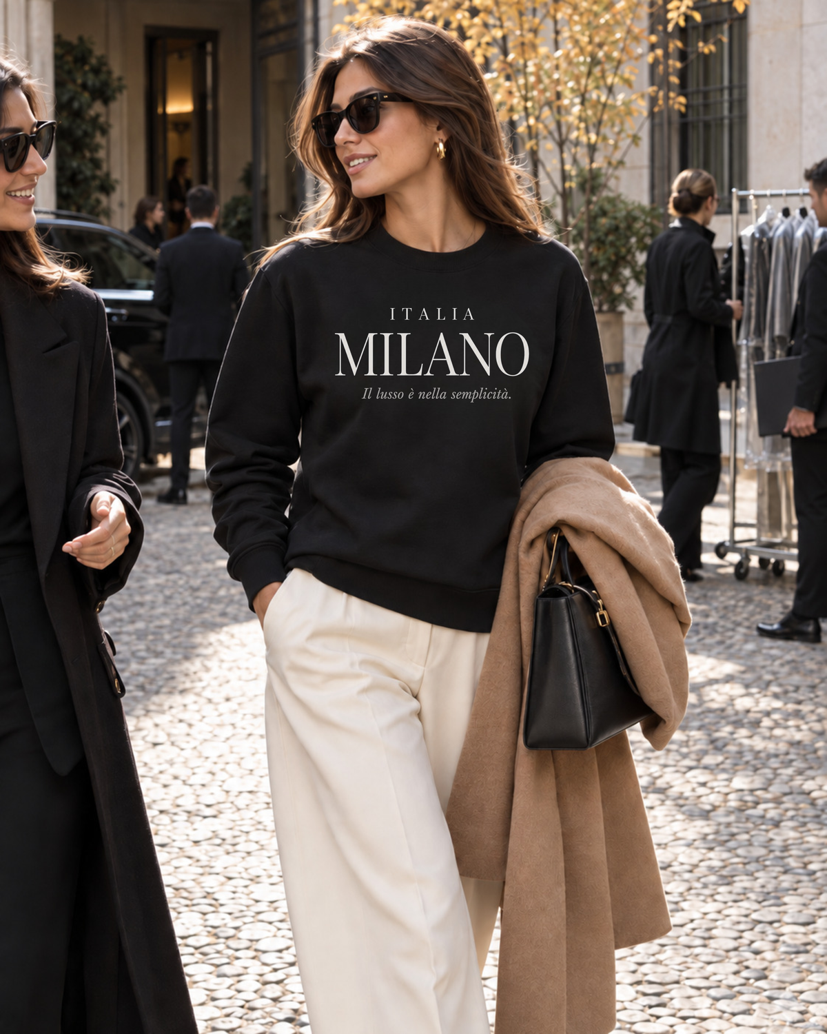 Woman wearing a black Milano Italy sweatshirt with cream tailored trousers and camel coat in an elegant Milan courtyard during autumn fashion week season.