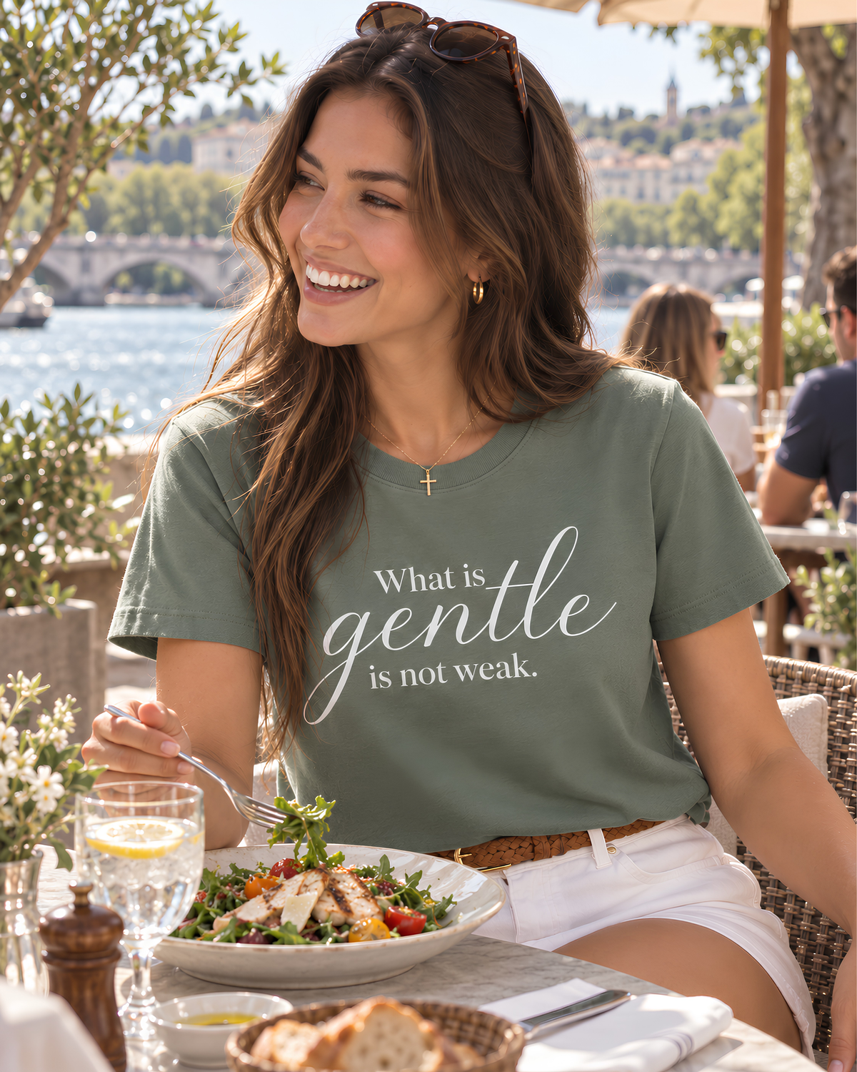 Young woman wearing a sage “What Is Gentle Is Not Weak” inspirational graphic tee with white shorts while having lunch at a bright riverside restaurant.