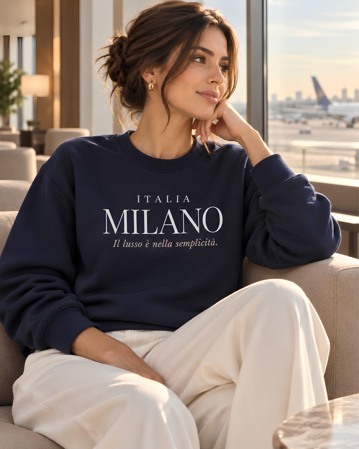 A woman sits in a beige armchair in a modern lounge setting, wearing a navy blue sweatshirt with 'ITALIA MILANO' printed in white across the chest, paired with cream-colored pants.