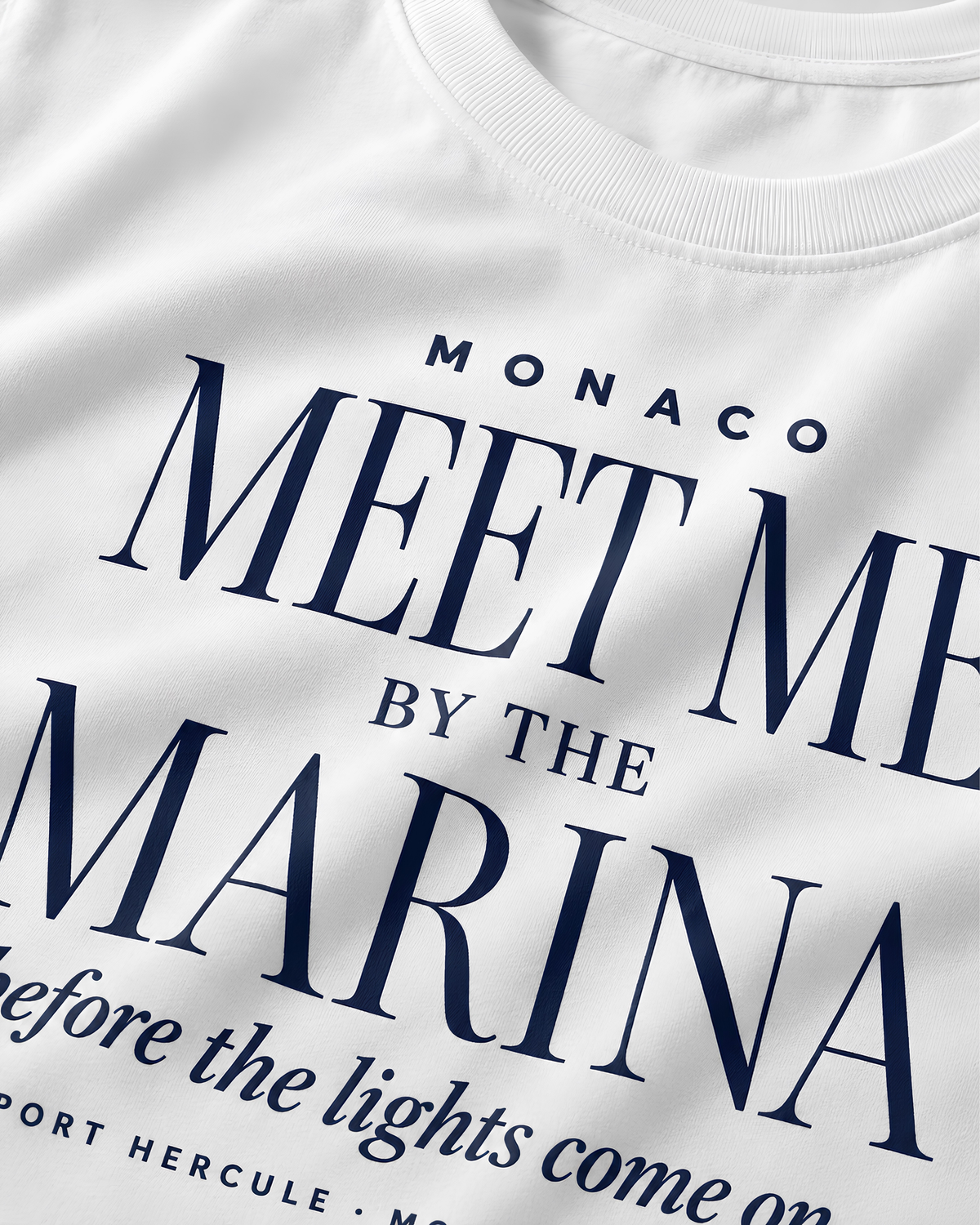 Close-up of a white crewneck t-shirt featuring navy blue printed text that reads 'MONACO MEET ME BY THE MARINA before the lights come on' and 'PORT HERCULE - MONACO'.
