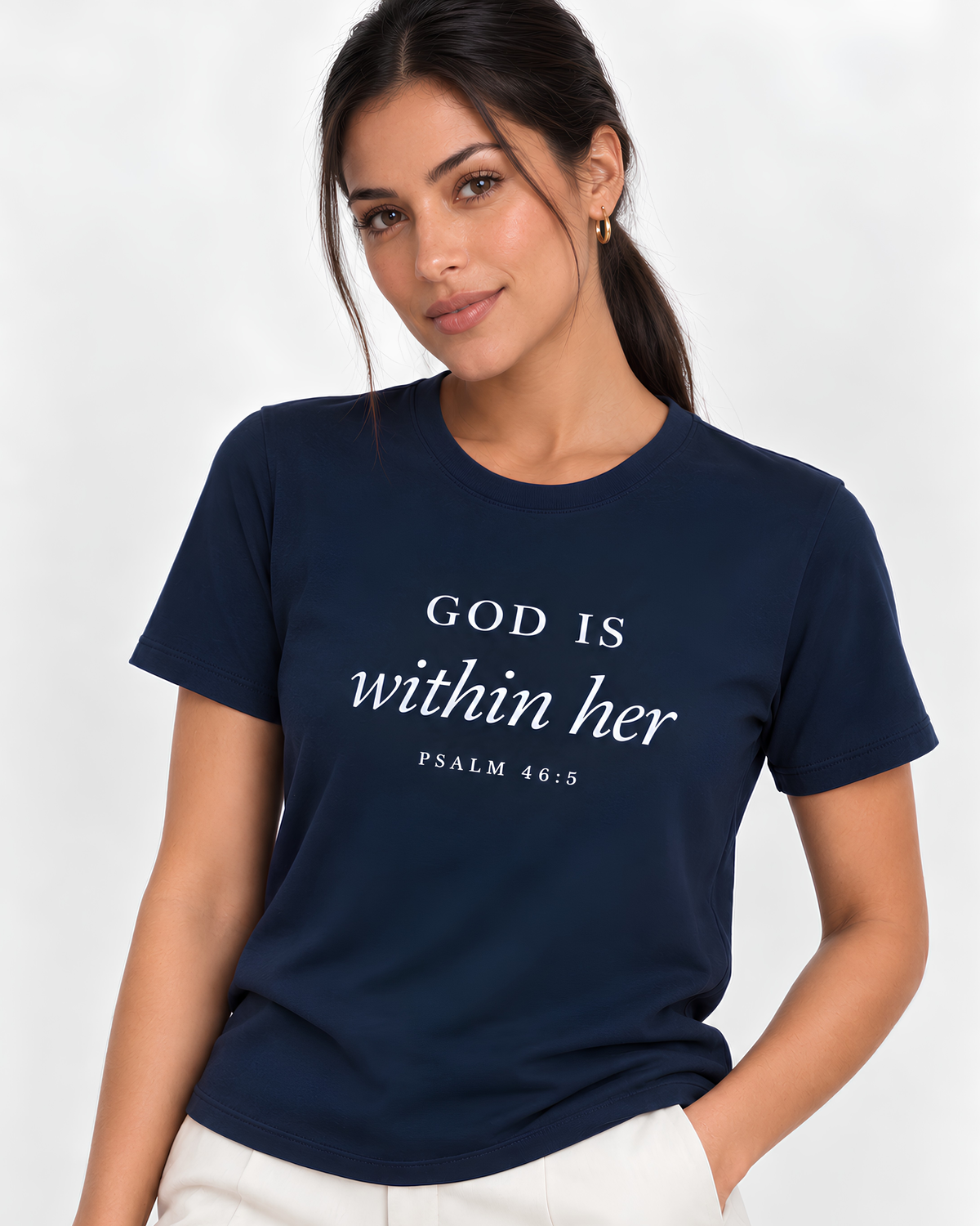 A woman wearing a navy blue crewneck t-shirt with the white printed text 'GOD IS within her PSALM 46:5' on the front, paired with off-white pants, against a light gray studio background.