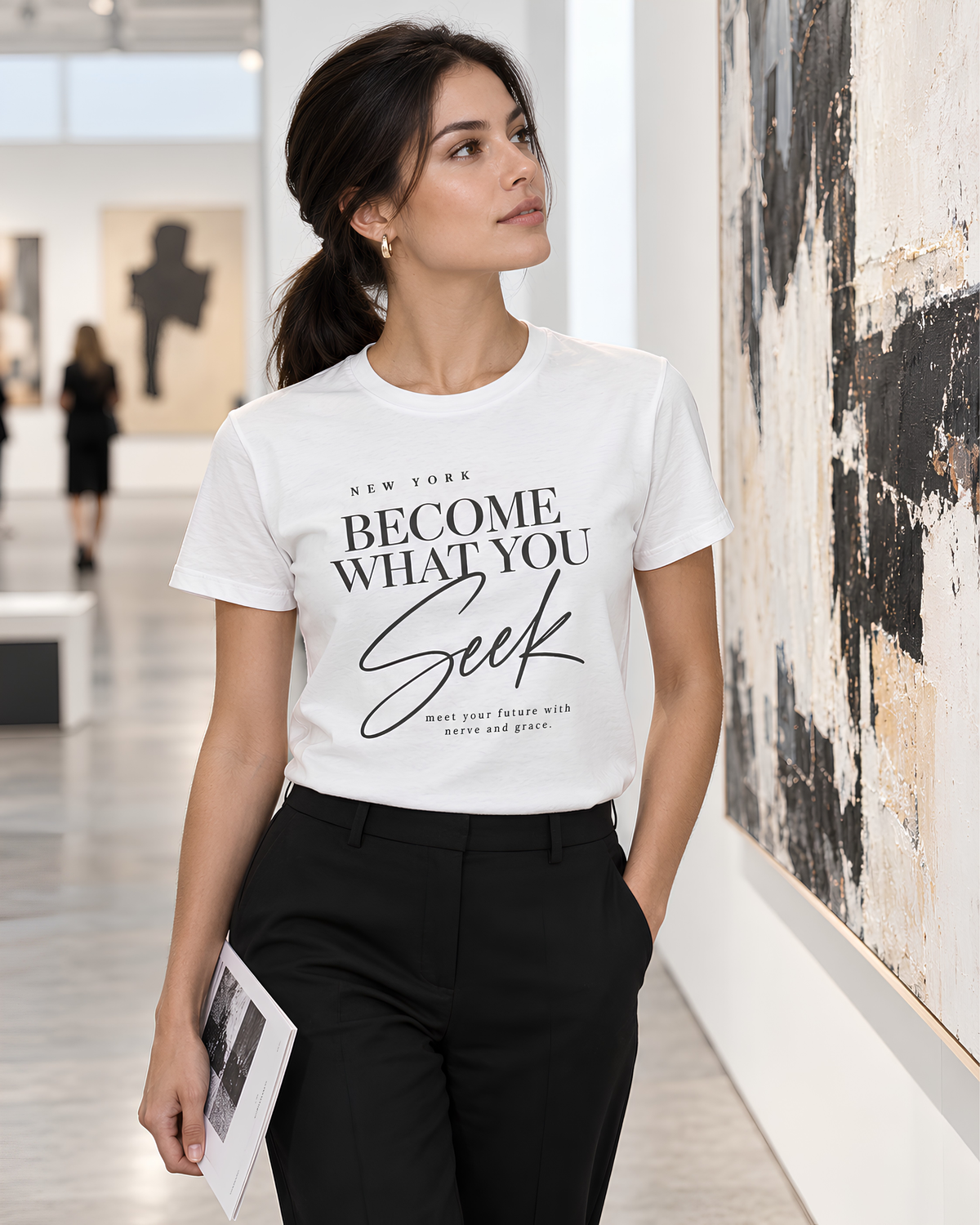 A woman wearing a white graphic T-shirt with the text 'BECOME WHAT YOU SEEK' in black script, paired with black pants, standing in a bright art gallery next to a large abstract painting.