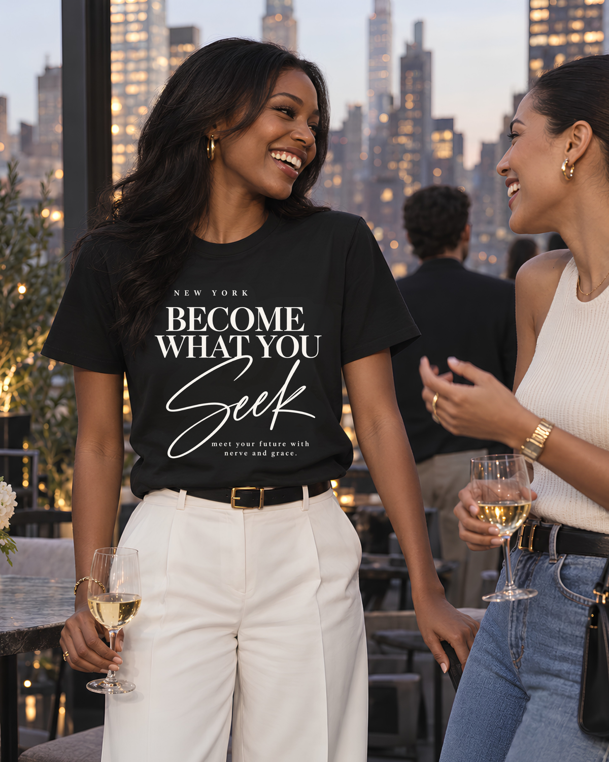 Black woman wearing a black New York graphic tee for women with “Become What You Seek” typography while enjoying a Manhattan rooftop evening with a friend.