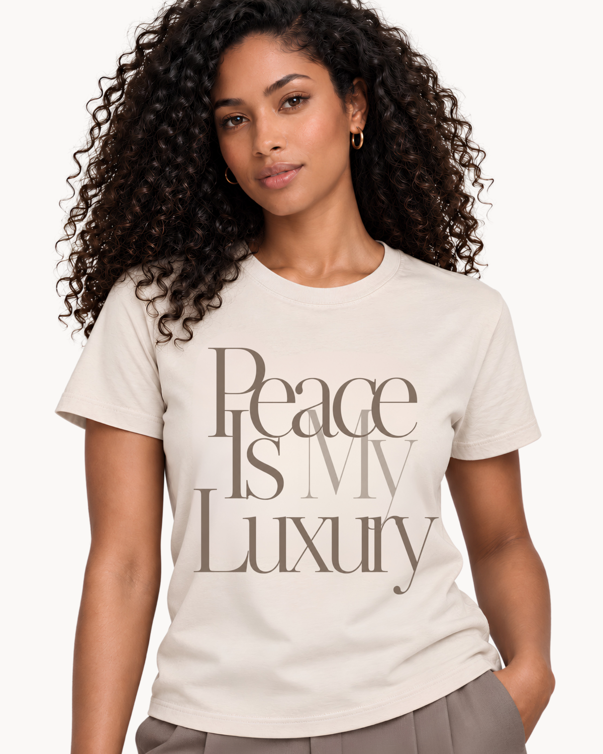 Woman wearing a soft bone T-shirt with oversized taupe serif typography reading “Peace Is My Luxury” against a clean light background.