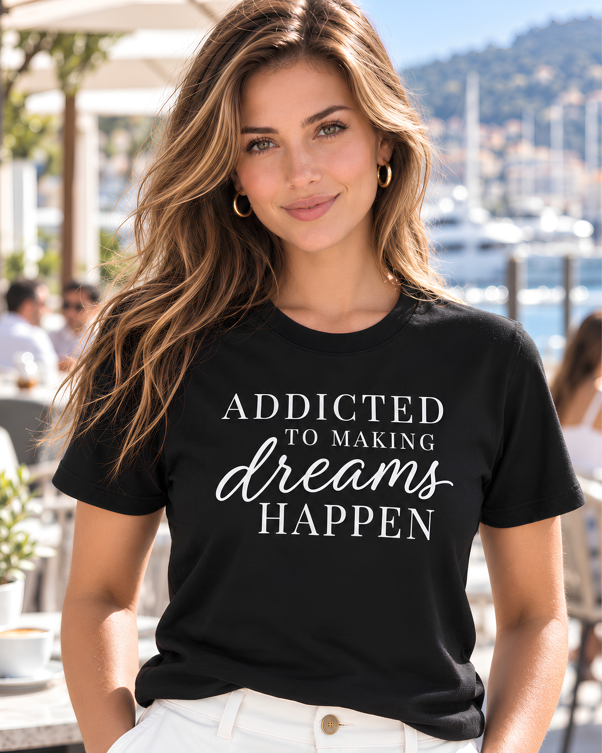 Young woman wearing a black Addicted to Making Dreams Happen motivational graphic tee with white shorts at a sunny marina café.