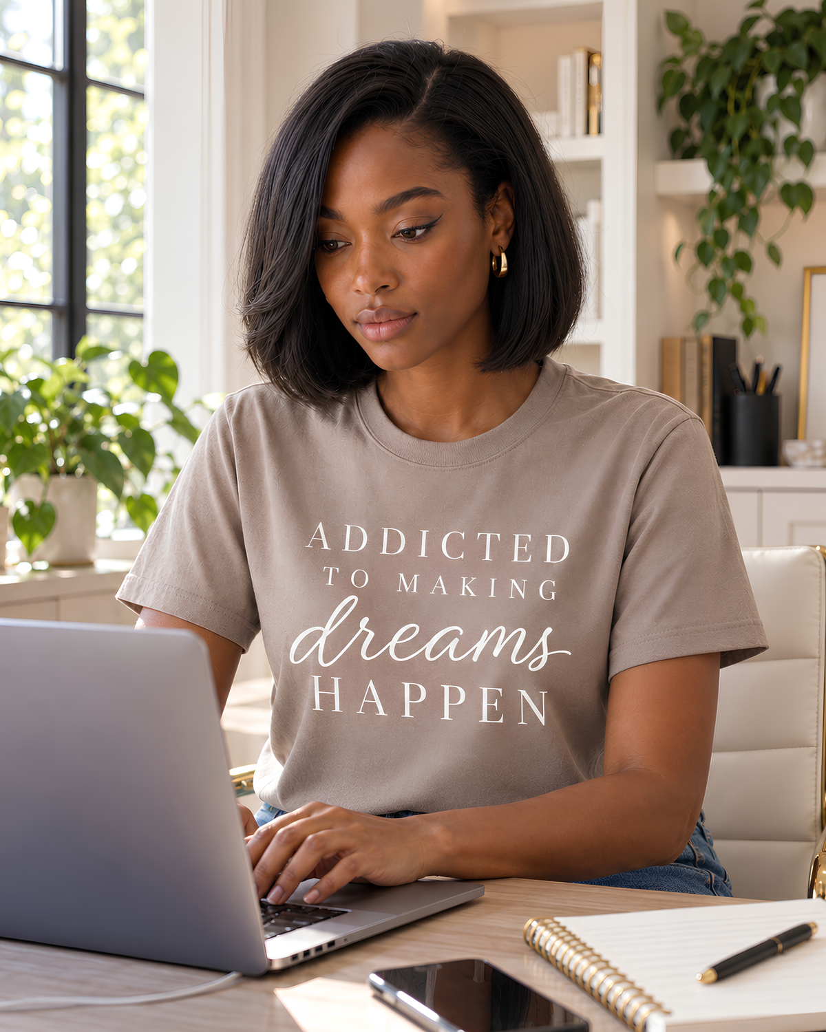 Woman wearing a mushroom Addicted to Making Dreams Happen graphic tee while working on a laptop in a bright, stylish home office.
