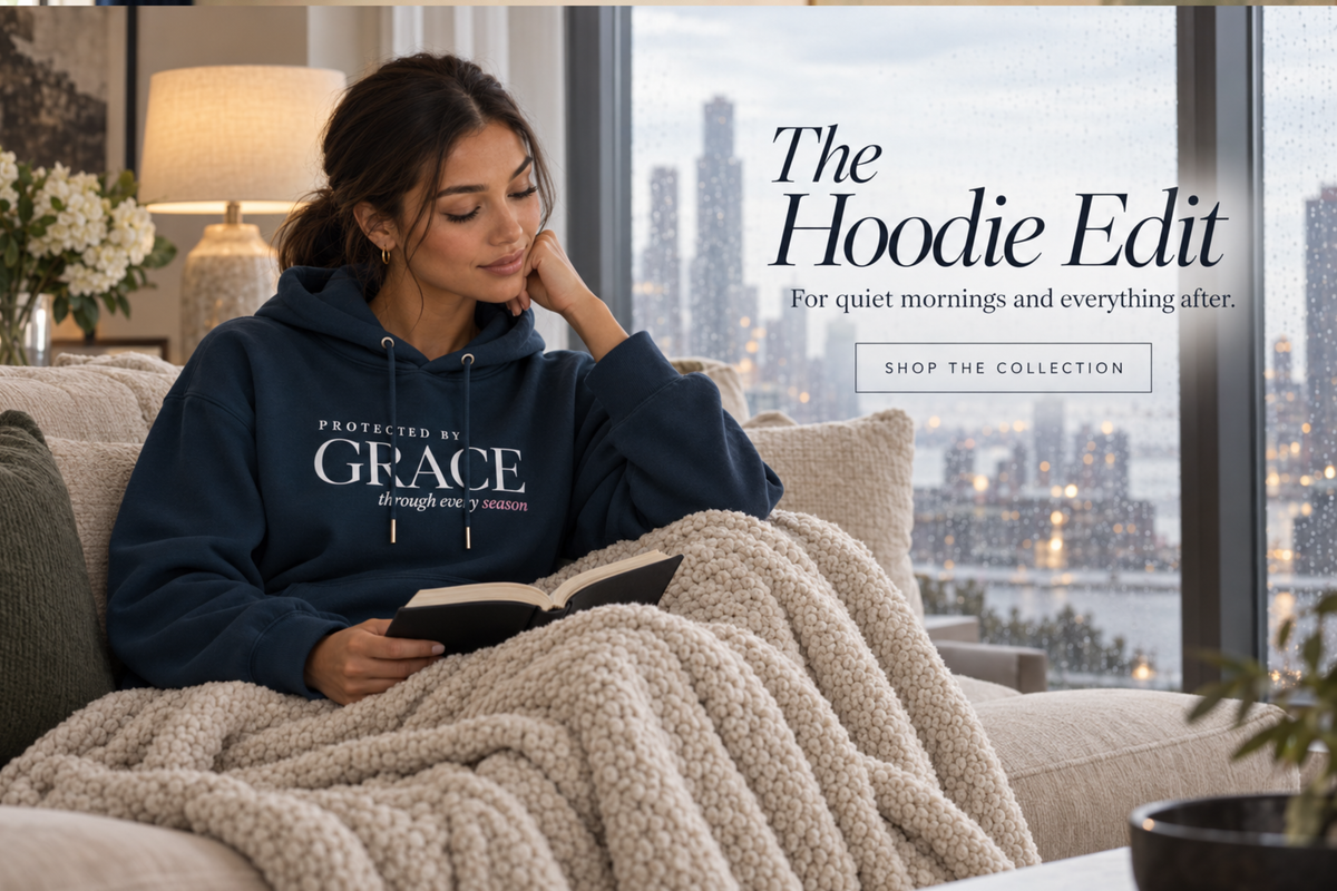 Navy Christian hoodie for women with “Protected by Grace through every season” faith graphic, styled in a luxury rainy-morning home as a woman reads the Bible under a cozy blanket.
