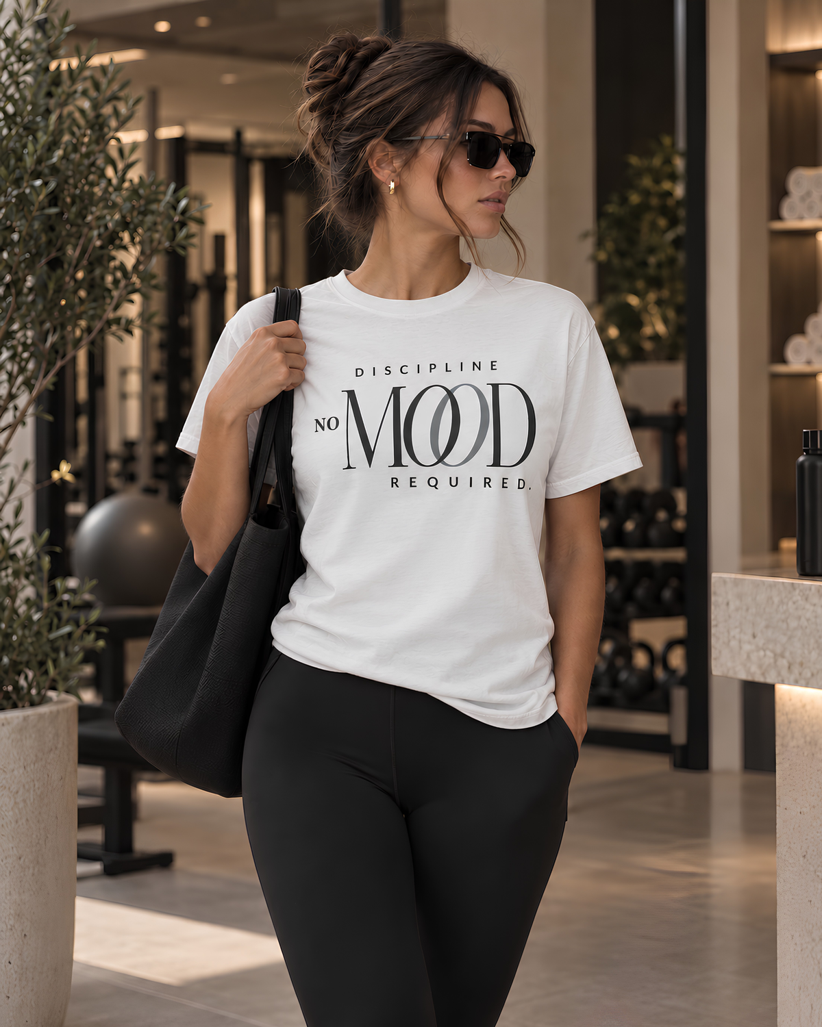 A woman wearing a white Pilates graphic t-shirt with the text 'DISCIPLINE NO MOOD REQUIRED' in black, paired with black leggings and carrying a black tote bag in a modern gym setting.