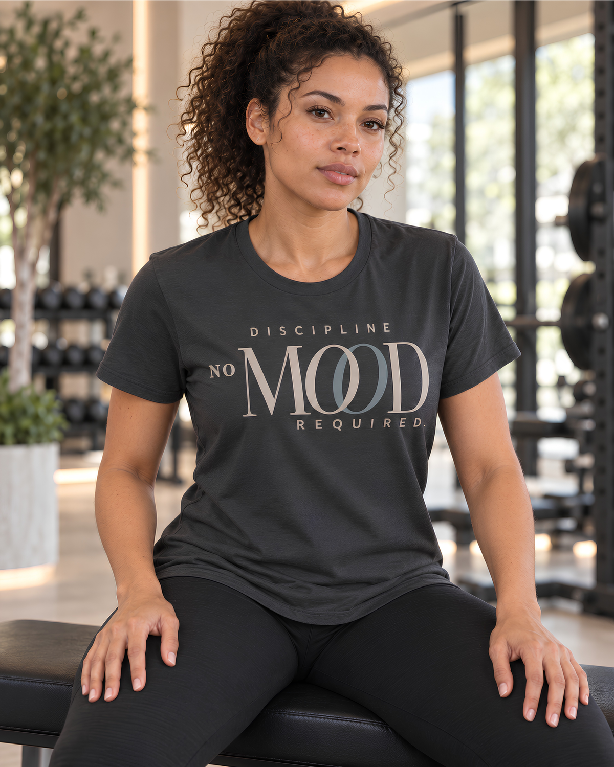 A woman with curly hair sits on a gym bench wearing a dark charcoal gray motivational graphic t-shirt with the text 'DISCIPLINE NO MOOD REQUIRED' in a metallic silver and gray font, paired with black leggings in a bright gym setting.