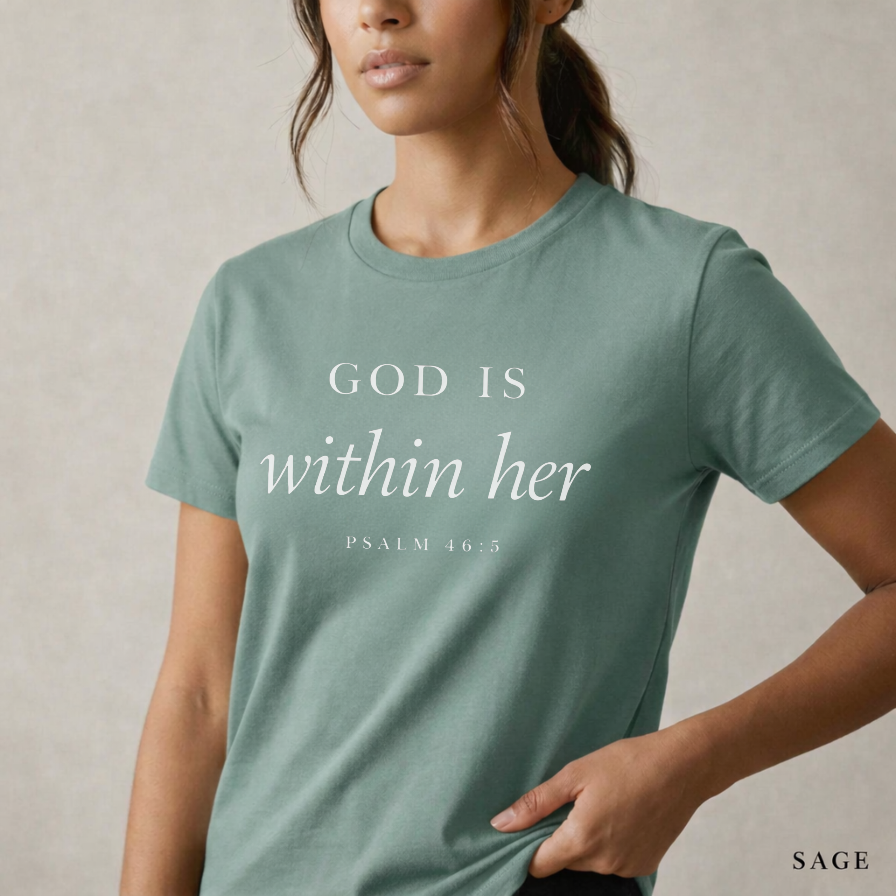 God is within her Psalm 46:5 bible verse on sage T-shirt. She is standing holding the scripture tee at the hem.