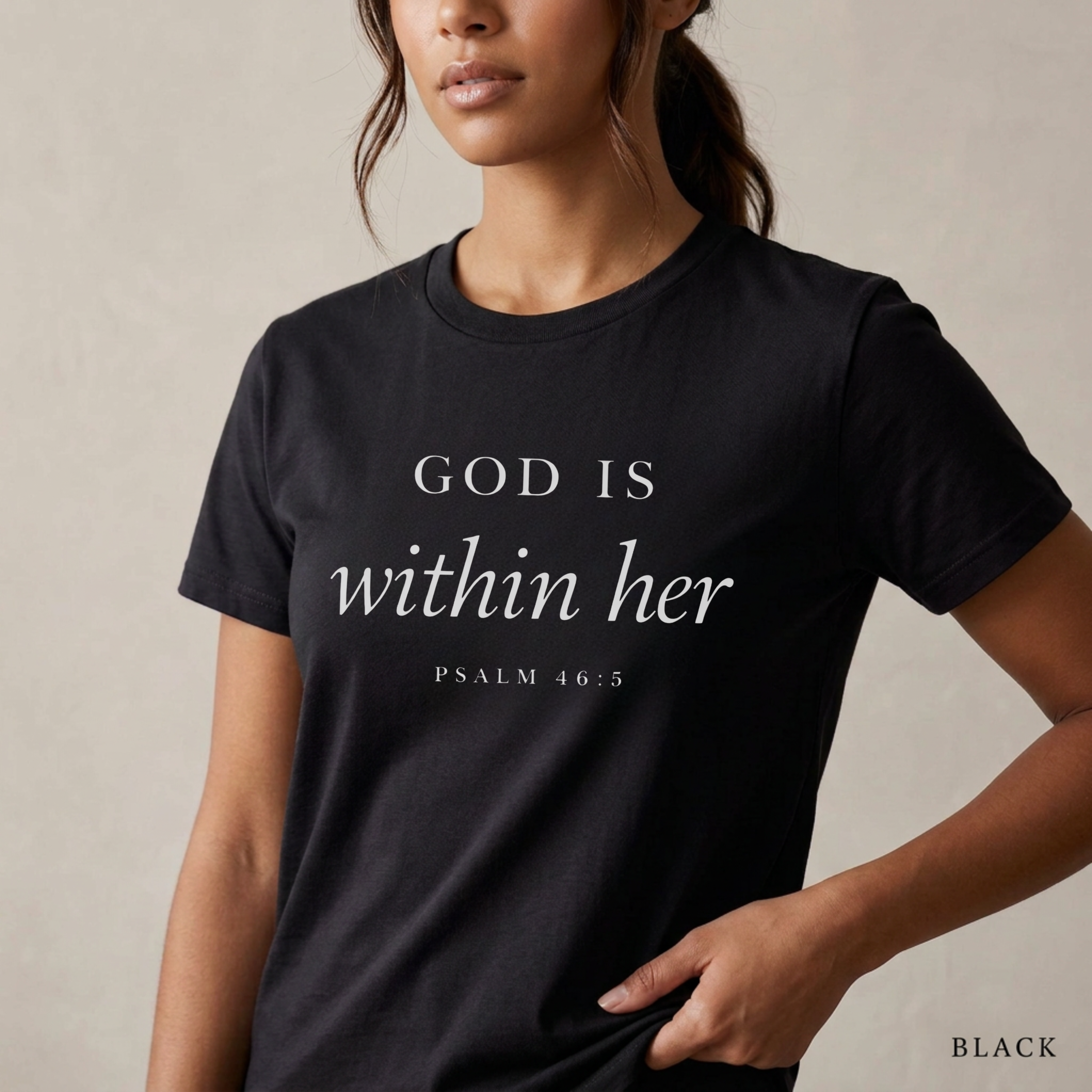 God is within her Psalm 46:5 bible verse on black T-shirt. She is standing holding the scripture tee at the hem.