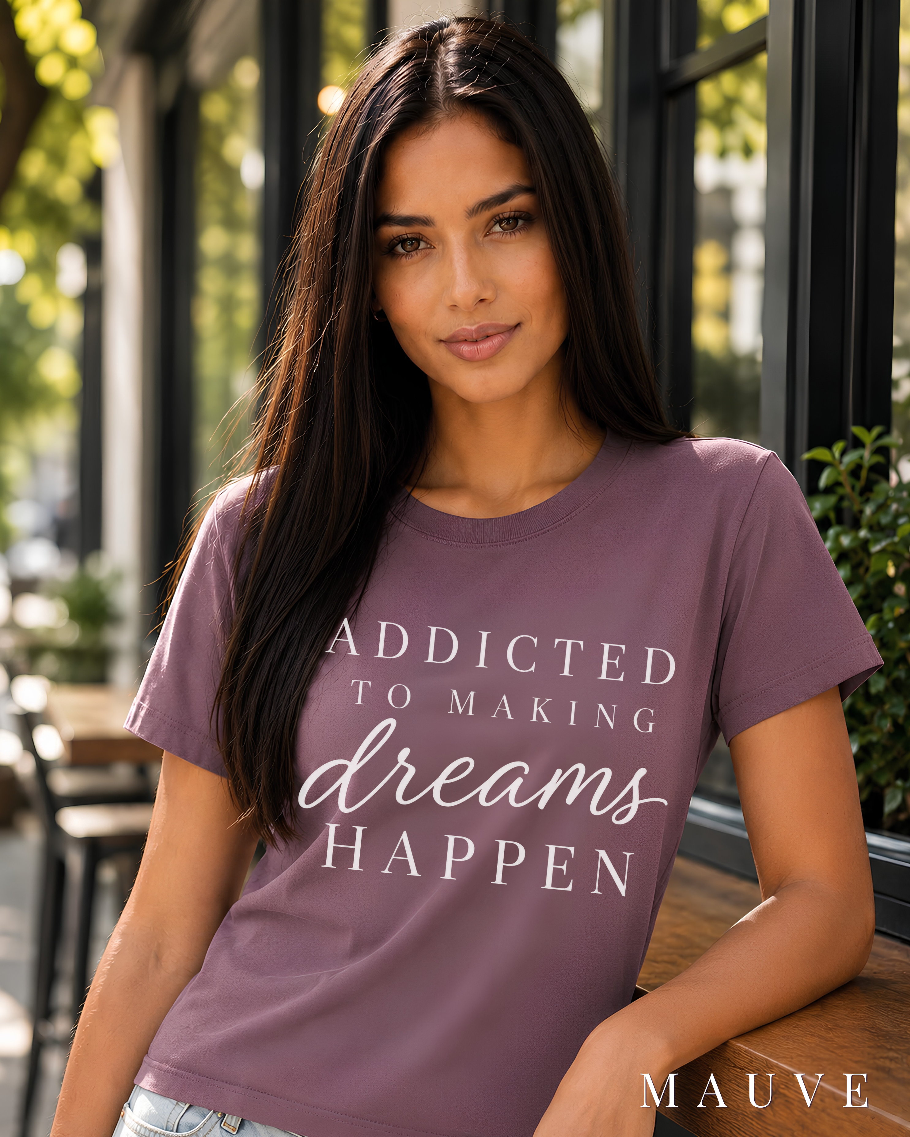 A woman wearing a mauve purple graphic t-shirt with the text 'ADDICTED TO MAKING dreams HAPPEN' in white, leaning on a wooden counter in a cafe setting.