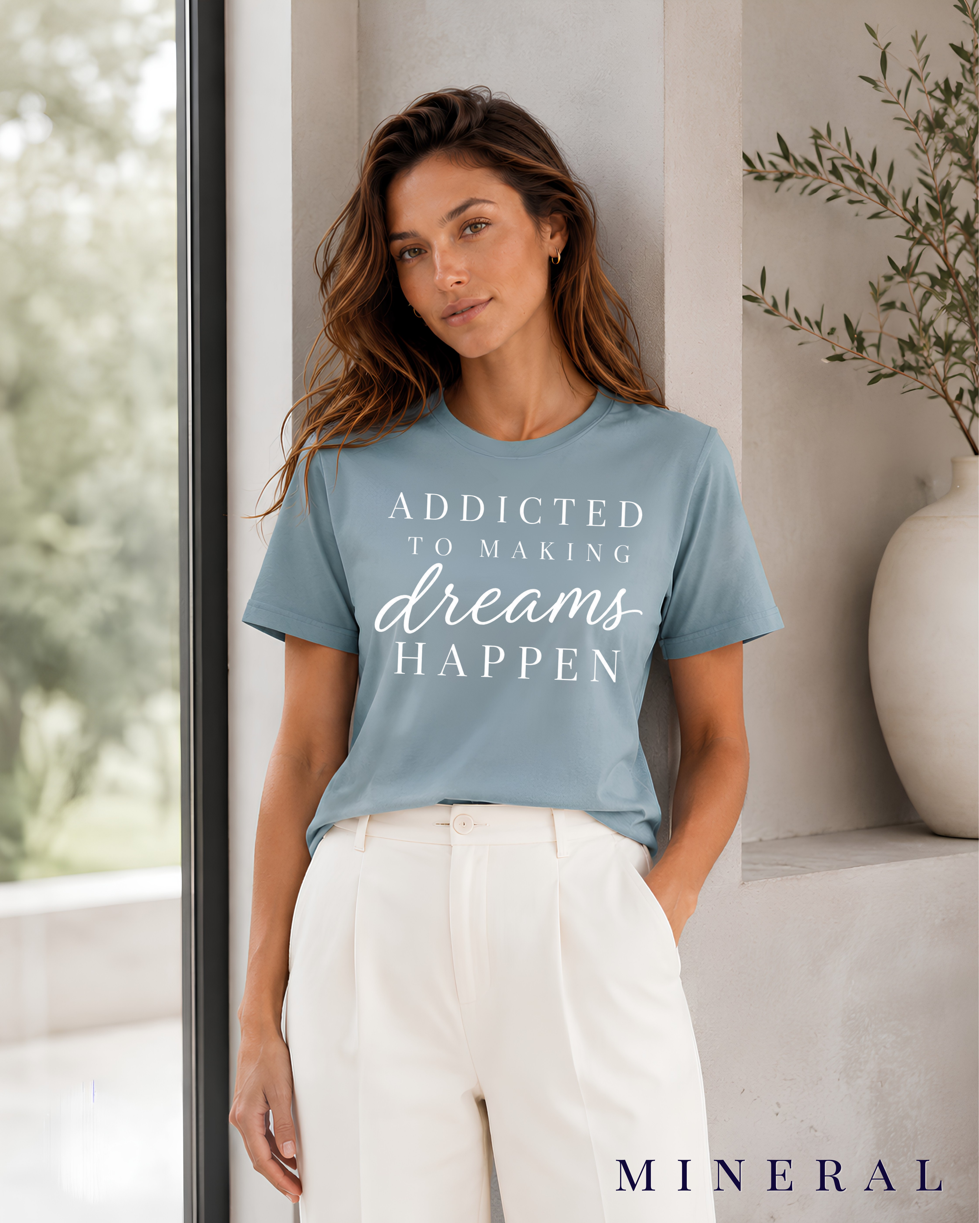 A woman wearing a mineral motivational graphic t-shirt with the text 'ADDICTED TO MAKING dreams HAPPEN' in white, paired with white trousers, standing in a modern interior setting with a large window and a white vase. Color label MINERAL in the bottom left corner.