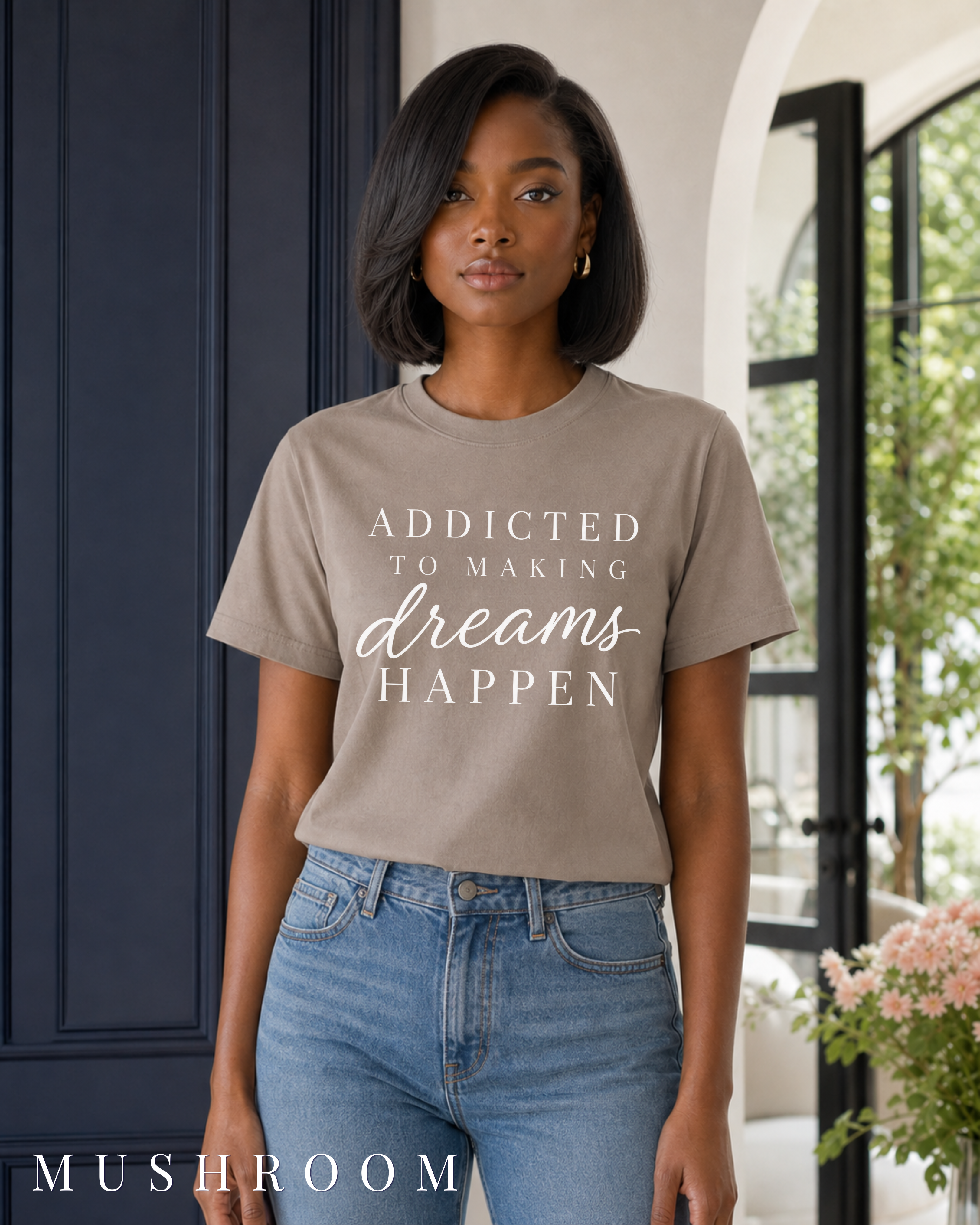 A woman wearing a motivational taupe graphic t-shirt with the text 'ADDICTED TO MAKING dreams HAPPEN' in white, paired with blue jeans, standing in a doorway with a dark navy paneled door on the left. Color label MUSHROOM in the bottom left corner.