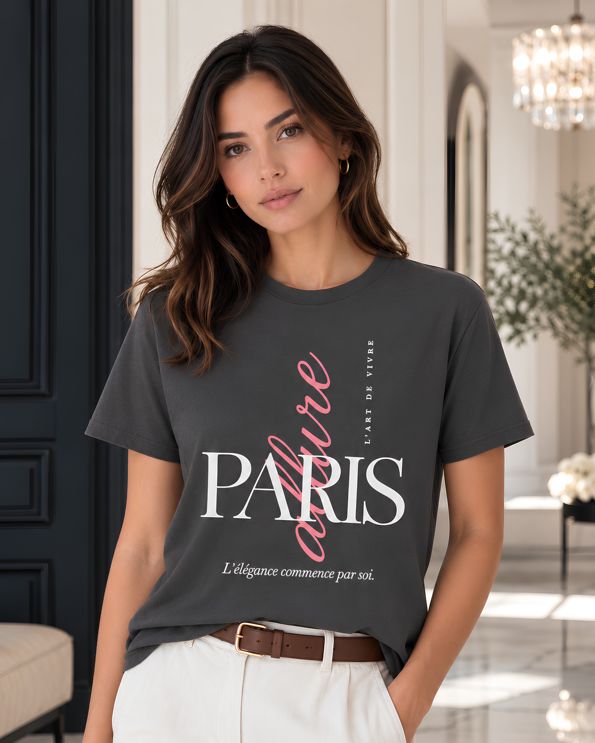 A woman wearing a dark gray graphic T-shirt with 'PARIS' and 'L'Art de Vivre' printed on the front, paired with white trousers, in a bright, elegant indoor setting.
