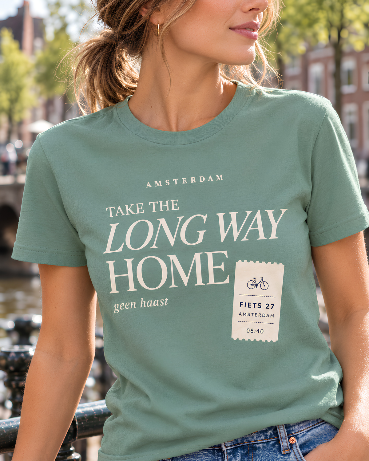 A woman wearing a sage green t-shirt with a white graphic print that reads 'AMSTERDAM TAKE THE LONG WAY HOME' and 'FIETS 27 AMSTERDAM', photographed outdoors in Amsterdam.