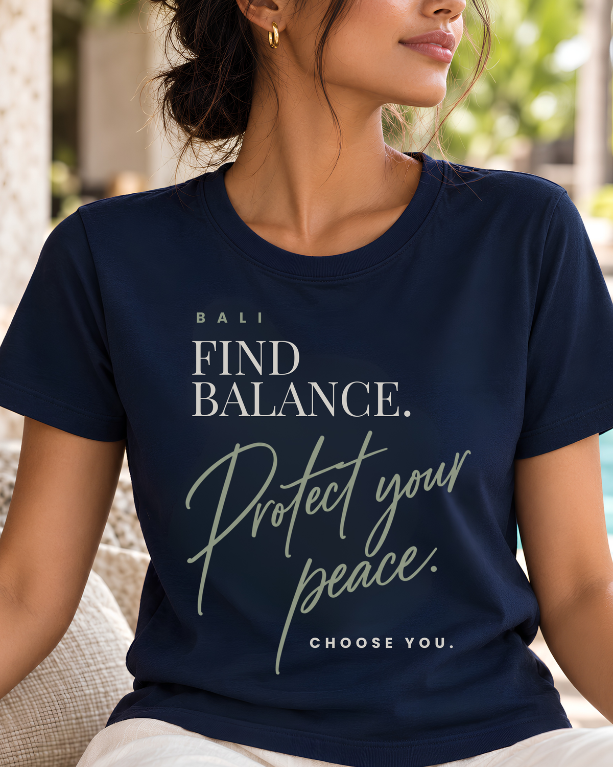 A woman wearing a navy blue graphic t-shirt with white and sage green text that reads 'BALI FIND BALANCE. Protect your peace.