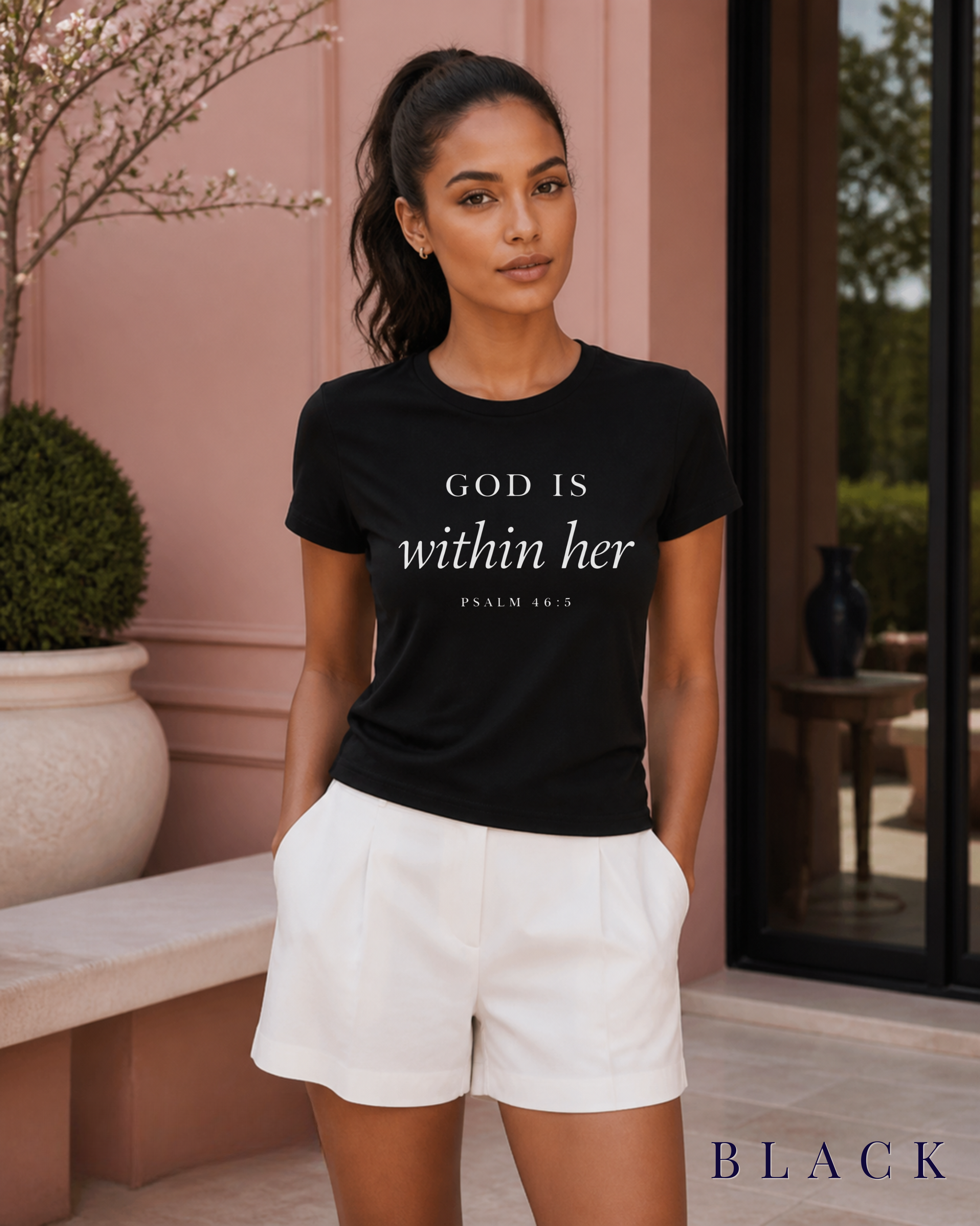 A woman wearing a black Christian graphic t-shirt with the text 'GOD IS within her' and white shorts, standing in front of a pink stucco building. Color label BLACK in bottom right corner.