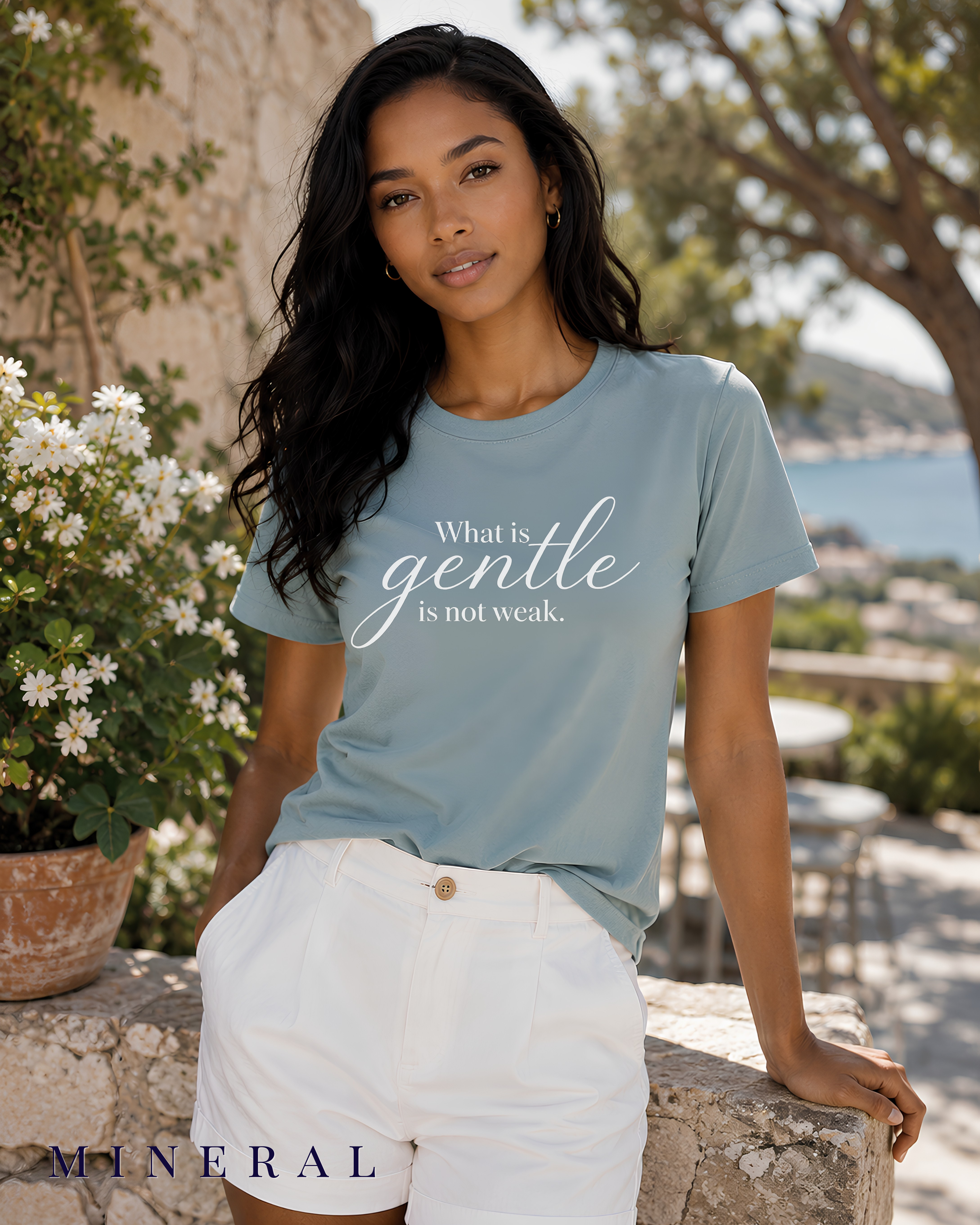 A woman wearing a mineral blue Christian graphic t-shirt with the text 'What is gentle is not weak.' in white script, paired with white shorts, posing outdoors on a stone ledge with a scenic background.