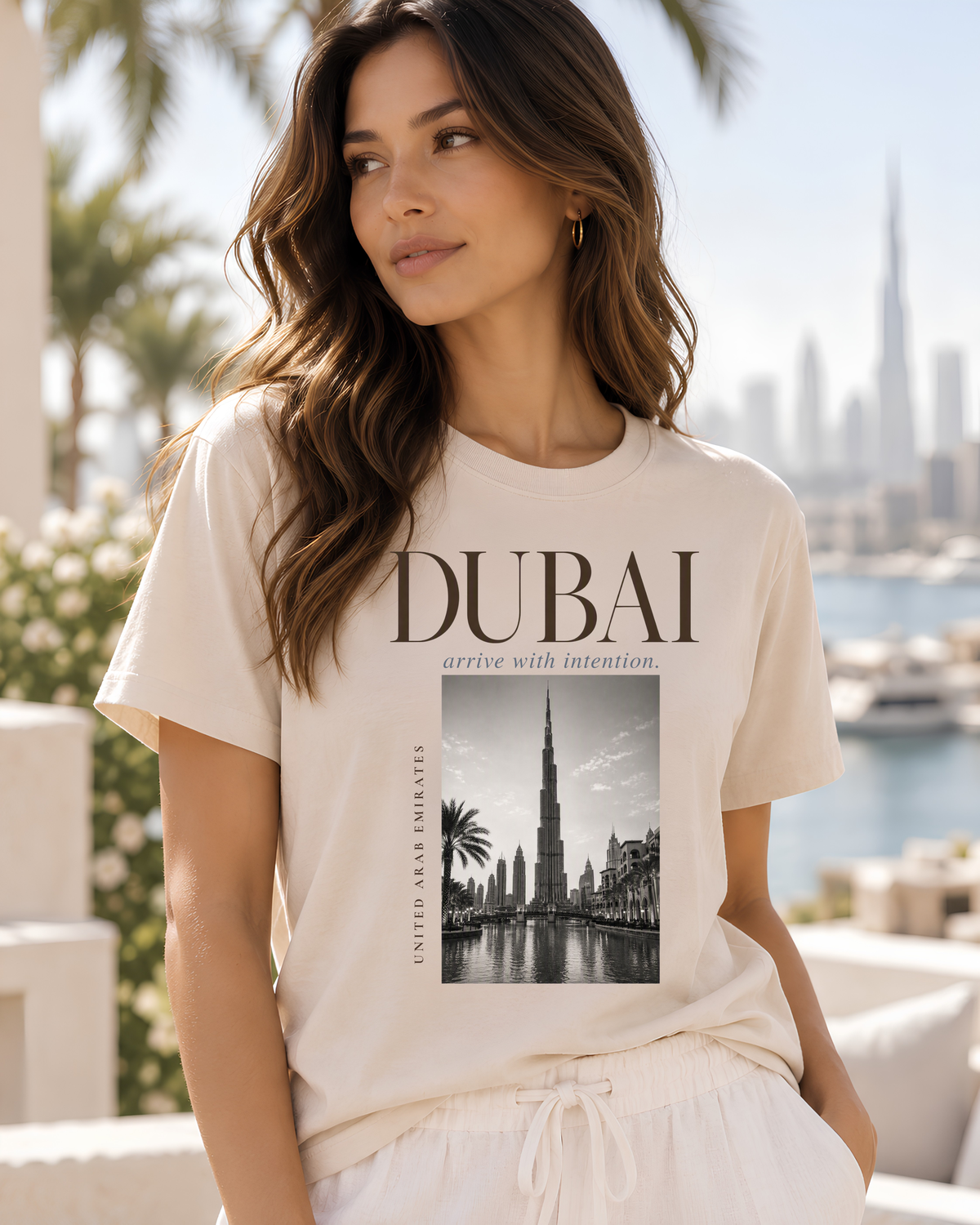 A woman wearing a cream-colored graphic t-shirt with 'DUBAI' and a Burj Khalifa print, set against a blurred background of a city skyline and water.