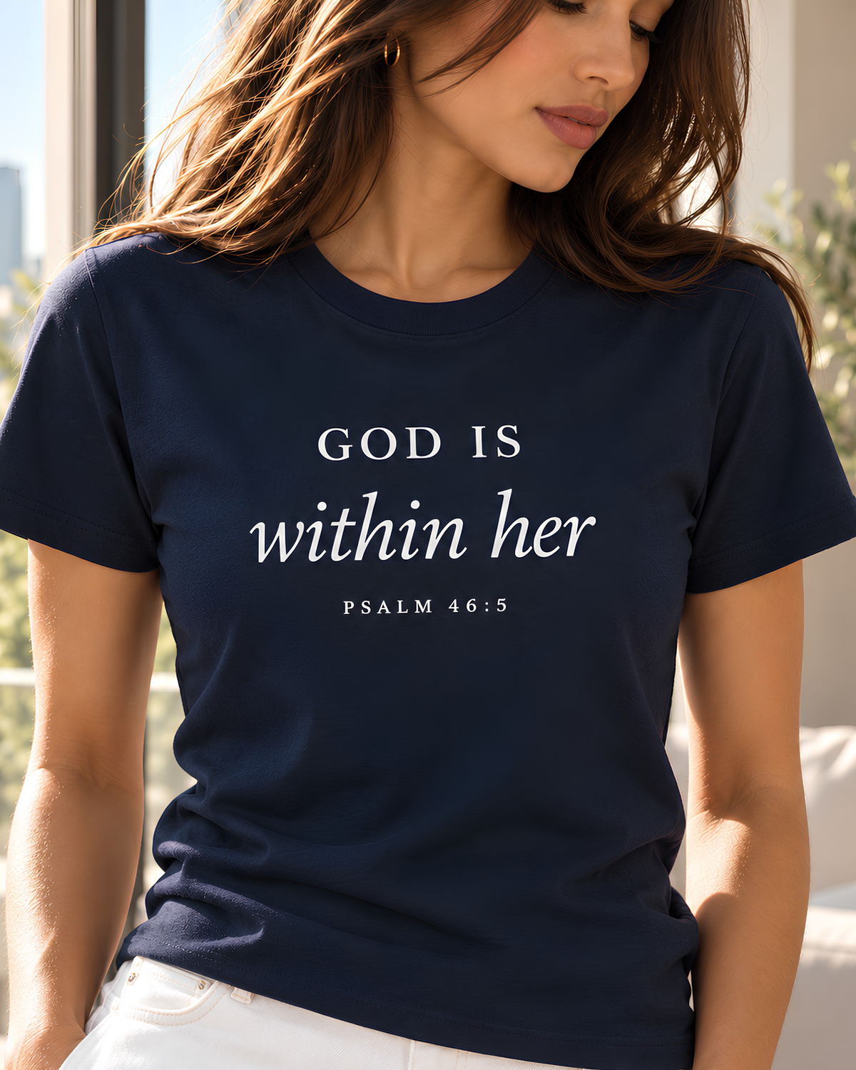 A woman wearing a navy blue crewneck t-shirt with the white printed text 'GOD IS within her PSALM 46:5' on the front, paired with off-white pants, against a light gray studio background.