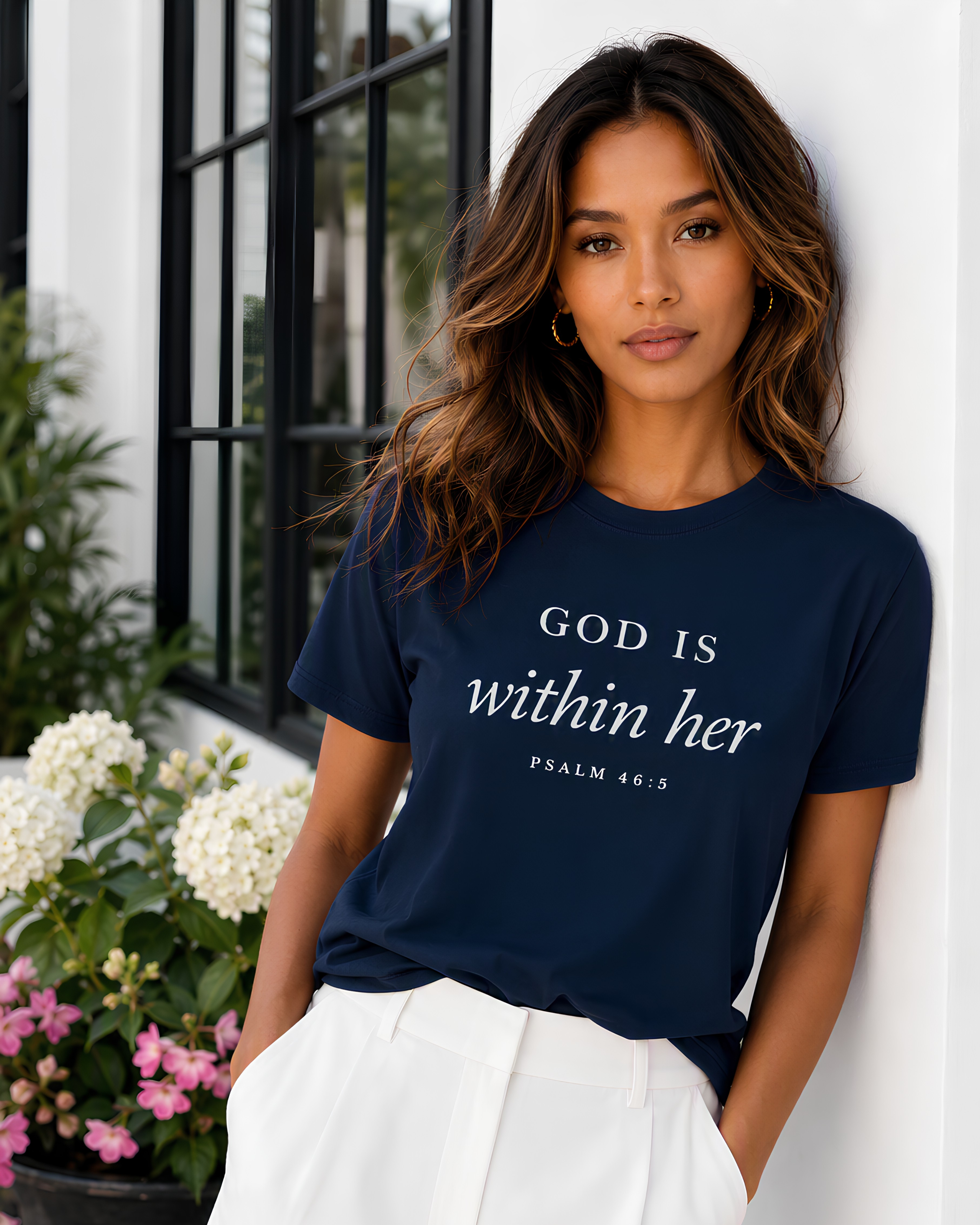 Woman wearing a navy God Is Within Her Psalm 46:5 Scripture tee paired with white pants, leaning against a white wall next to a window and flowers.