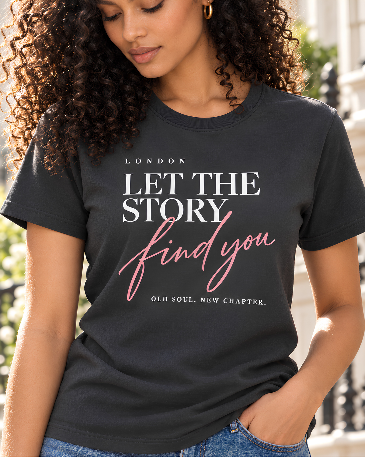 A woman with curly dark hair wearing a charcoal gray graphic t-shirt with the text 'LET THE STORY find you' in white and pink, paired with light beige trousers, against a light gray studio background.