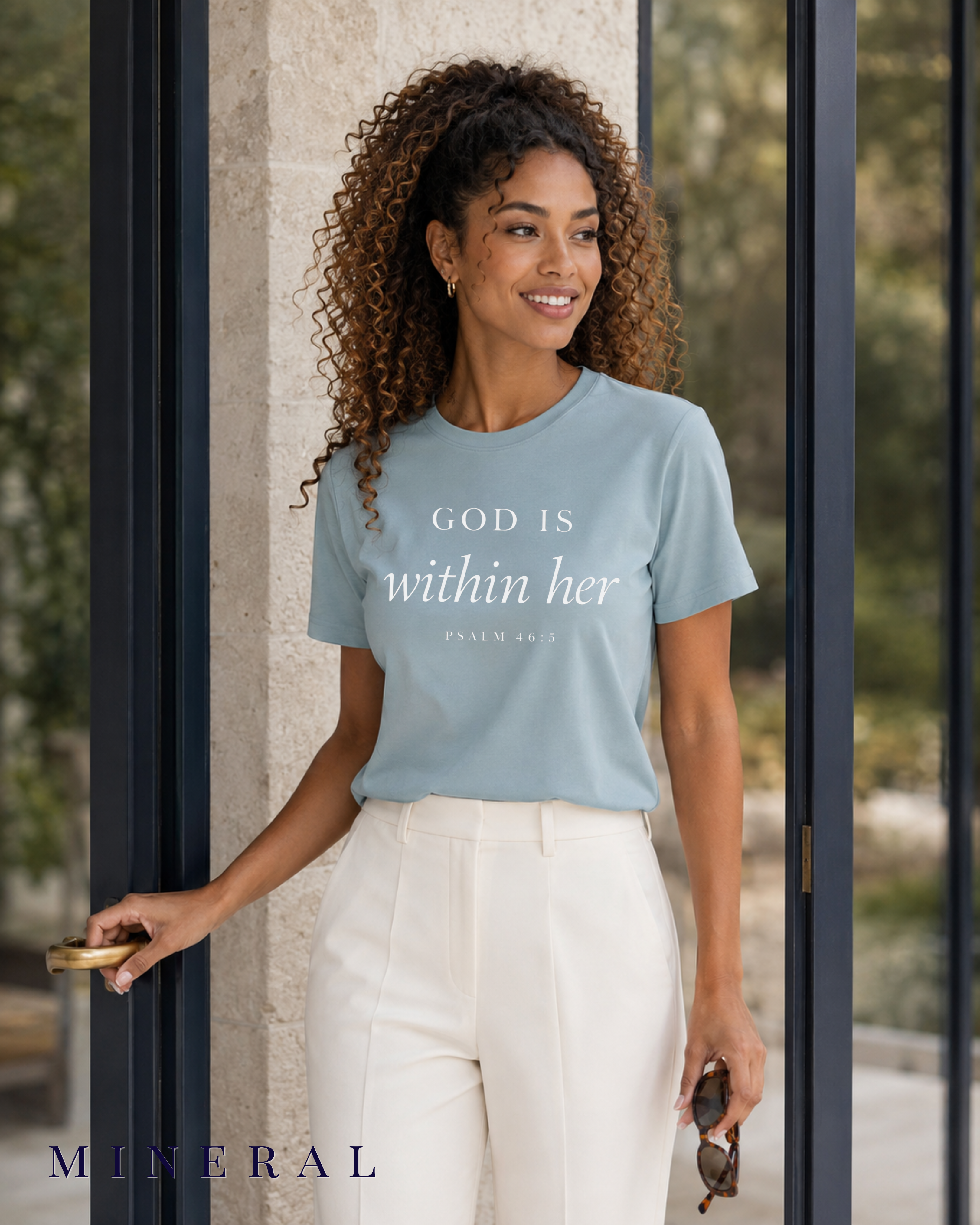 A woman wearing a light blue crewneck t-shirt with the text 'GOD IS within her' and 'PSALM 34:3' printed in white, paired with white pants, standing in front of a modern glass door. The color label 'MINERAL' is visible in the bottom left corner.