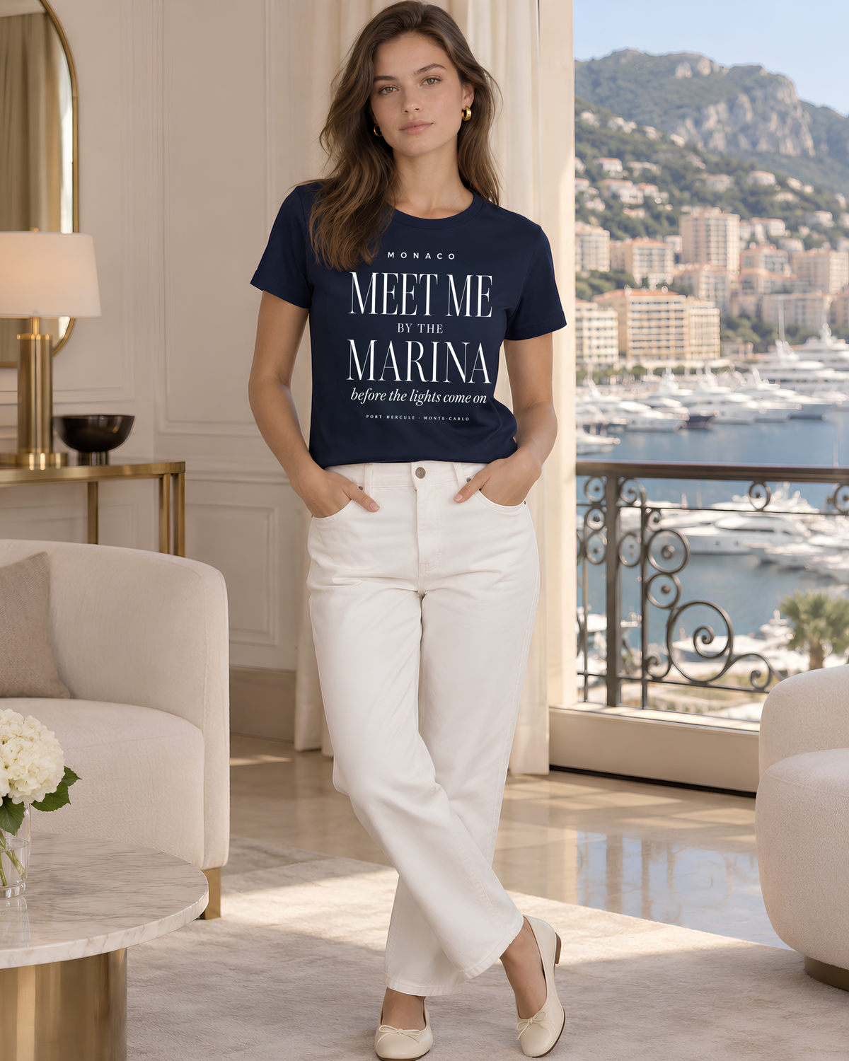 A woman stands in a luxurious interior setting with a balcony overlooking a marina and mountains, wearing a navy blue graphic t-shirt that reads 'MEET ME, BY THE MARINA' and white trousers.