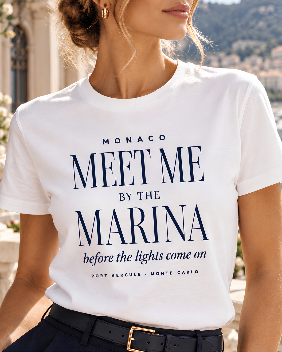 A woman wearing a white short-sleeve T-shirt with navy blue text that reads 'MONACO MEET ME BY THE MARINA before the lights come on' and 'FORT HERCULE - MONTE-CARLO'. The photo is cropped from the neck to the hips, with a blurred outdoor background.