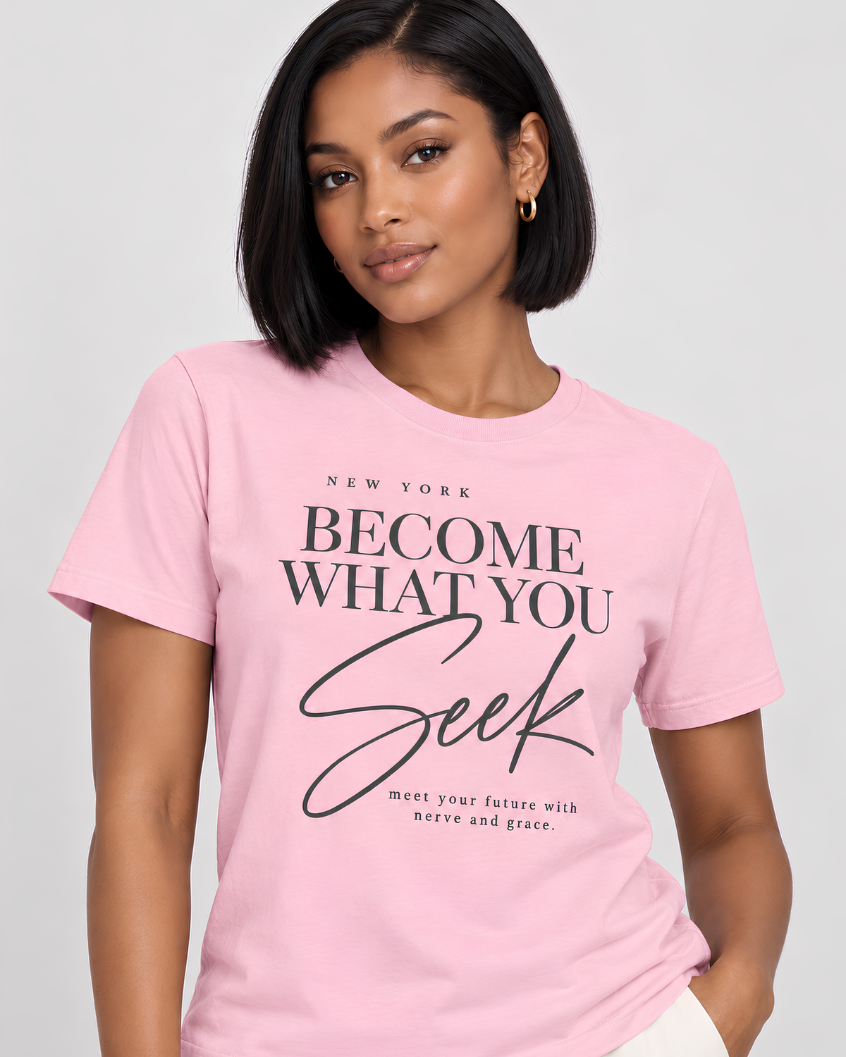A woman wearing a light pink short-sleeve graphic T-shirt with the text 'NEW YORK BECOME WHAT YOU Seek meet your future with nerve and gear.' printed in black on the front, photographed against a light gray studio background.
