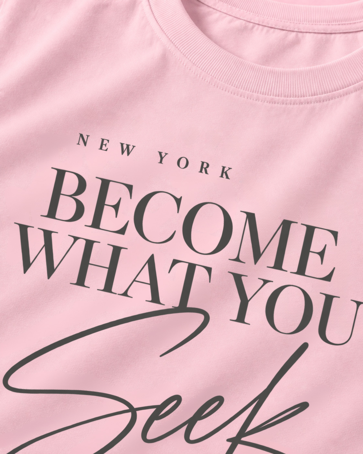 A close-up of a light pink crewneck t-shirt featuring the text 'NEW YORK BECOME WHAT YOU SEEK' printed in black.
