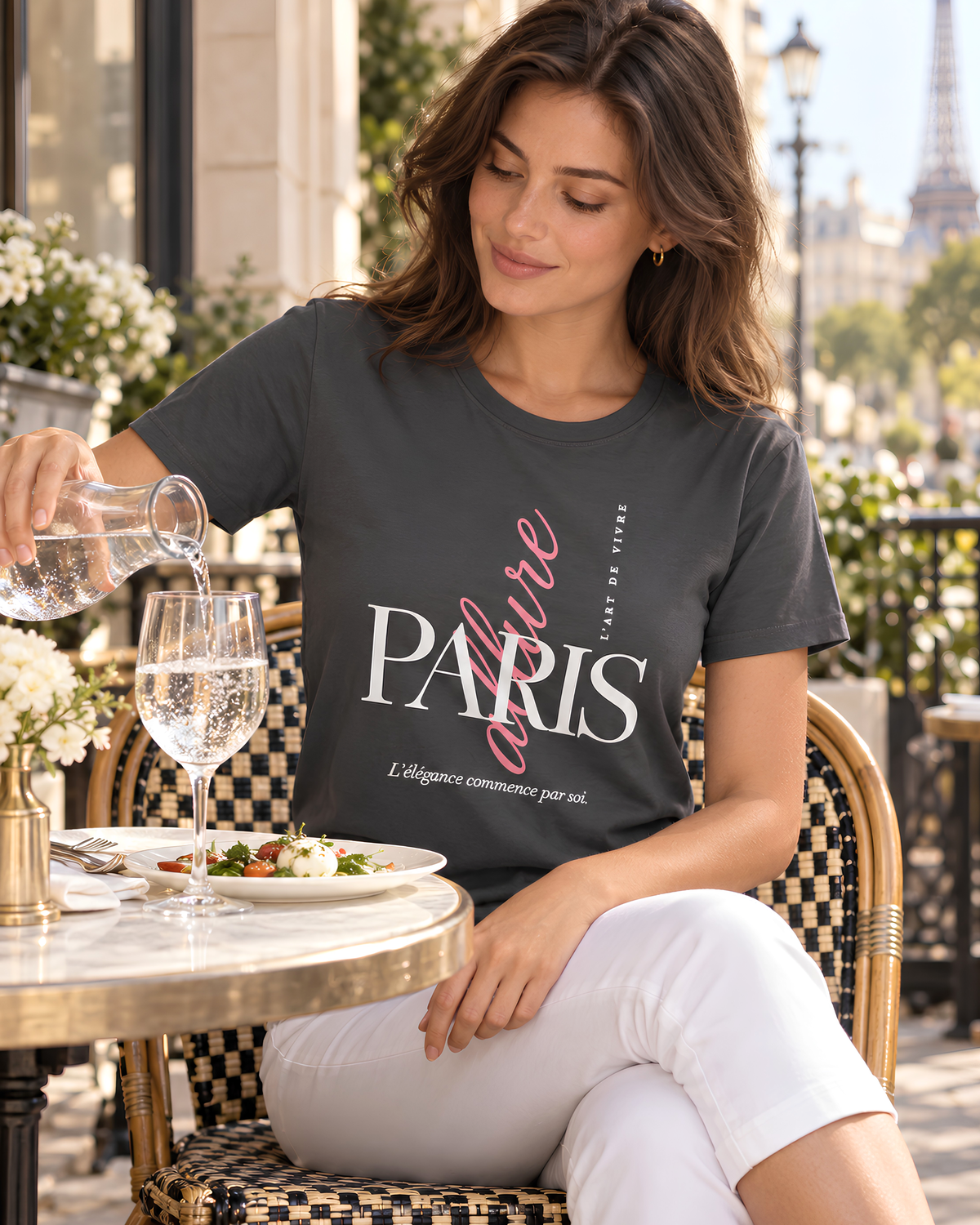 A woman sitting at an outdoor Parisian cafe table wearing a dark gray graphic t-shirt that reads 'Allure PARIS' in white and pink script, pouring water from a glass bottle into a wine glass.