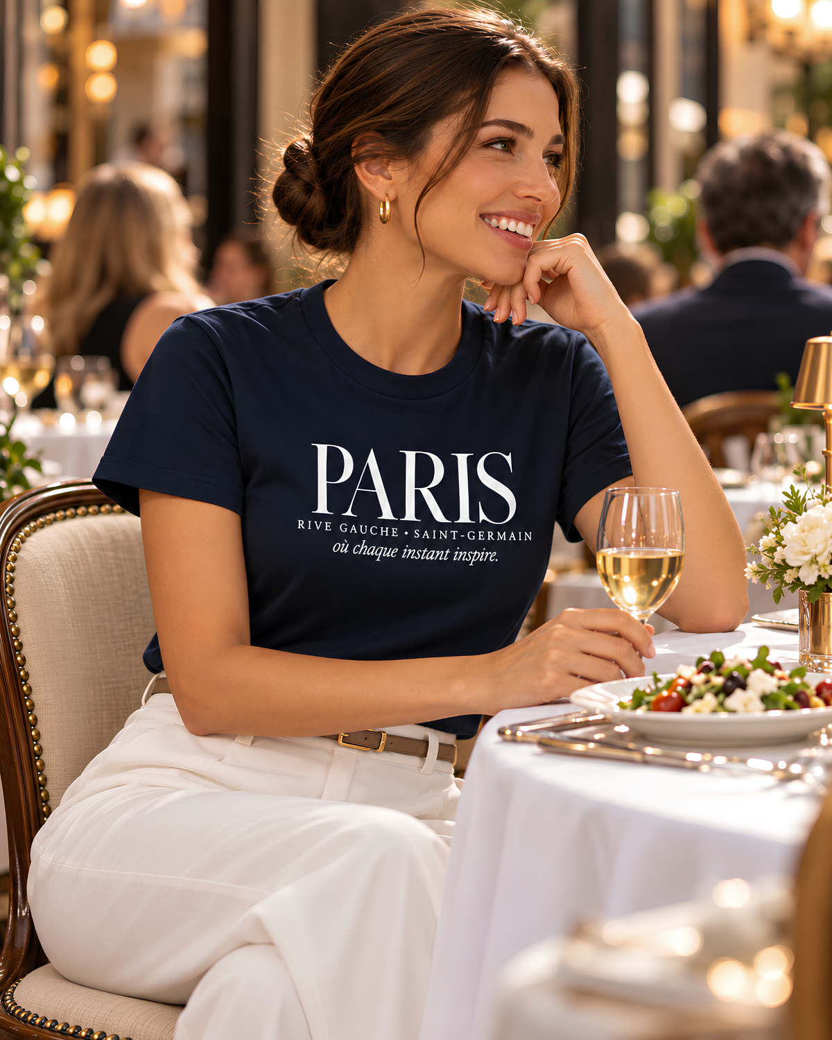 A woman seated at a restaurant table wearing a navy blue graphic T-shirt with white text reading 'PARIS RIVE GAUCHE-SAINT-GERMAIN en Champs-Élysées inspiré.' She is also wearing white pants, with a glass of white wine in front of her.