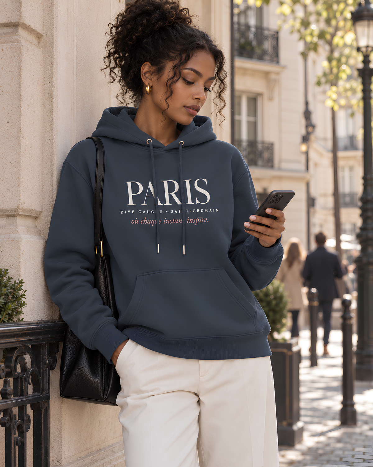A woman wearing a navy blue hoodie with 'PARIS' printed in white across the chest, standing outdoors on a city street and looking at her phone.