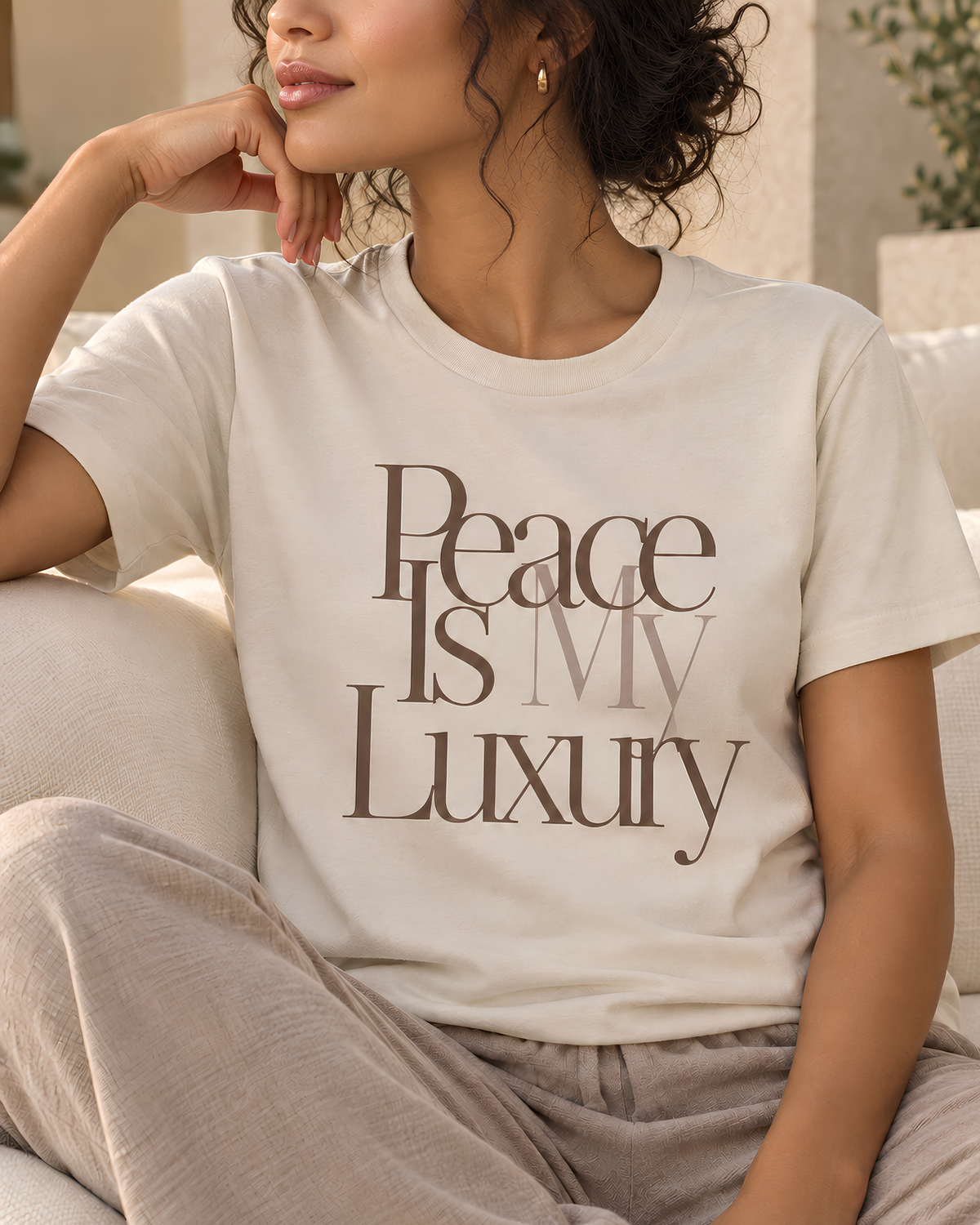 A woman wearing a cream-colored t-shirt with the text 'Peace Is My Luxury' in brown and gray lettering, paired with light gray lounge pants, sitting on a beige sofa.